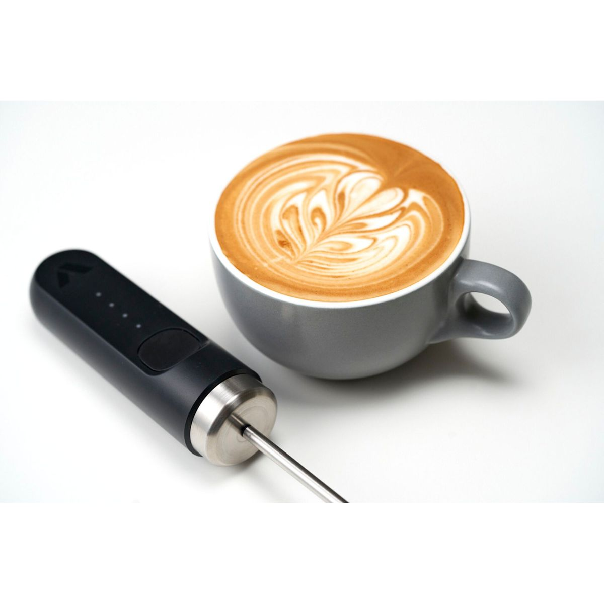 Eine graue Tasse Latte mit Herzmuster steht neben einem schwarzen Milchaufschäumer auf einer weißen Oberfläche.