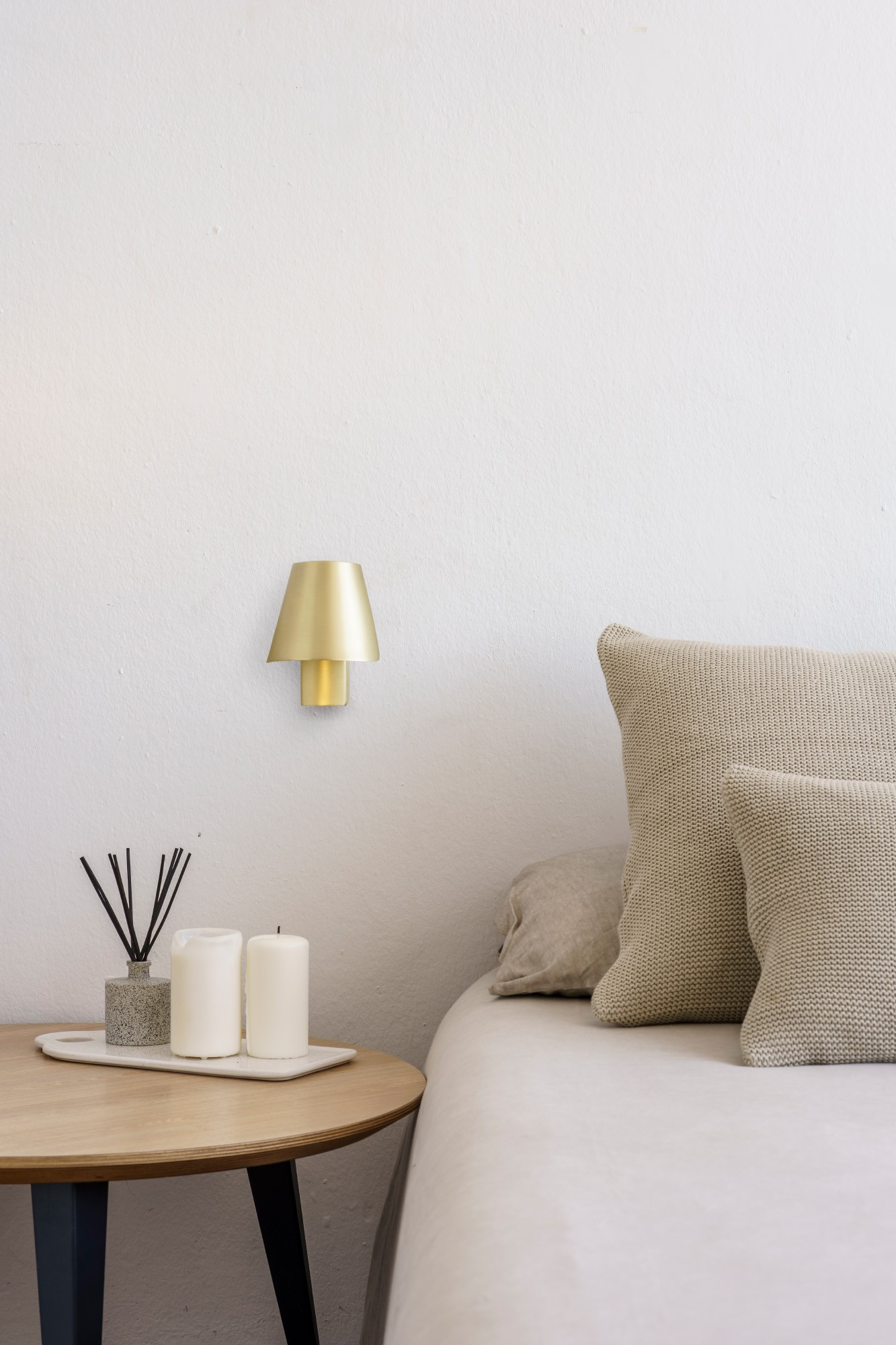 Scena camera da letto con tavolo rotondo, candele e lampada da parete dorata. Cuscini beige su letto contro muro bianco.