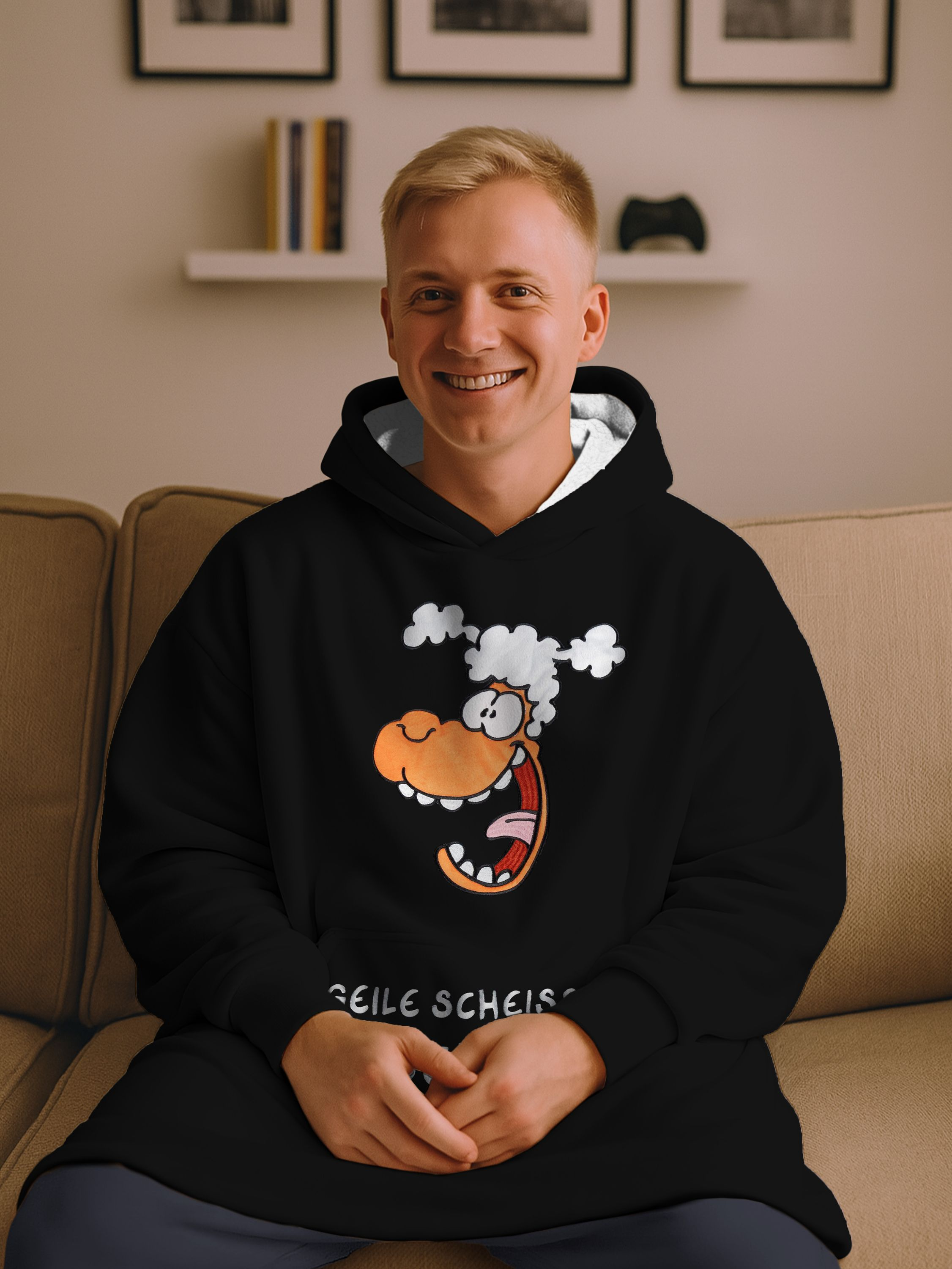 Ein lächelnder Mann trägt einen schwarzen Kapuzenpullover mit einem Cartoon-Gesicht. Er sitzt auf einer Couch. Im Hintergrund sind Bilder und ein Regal.