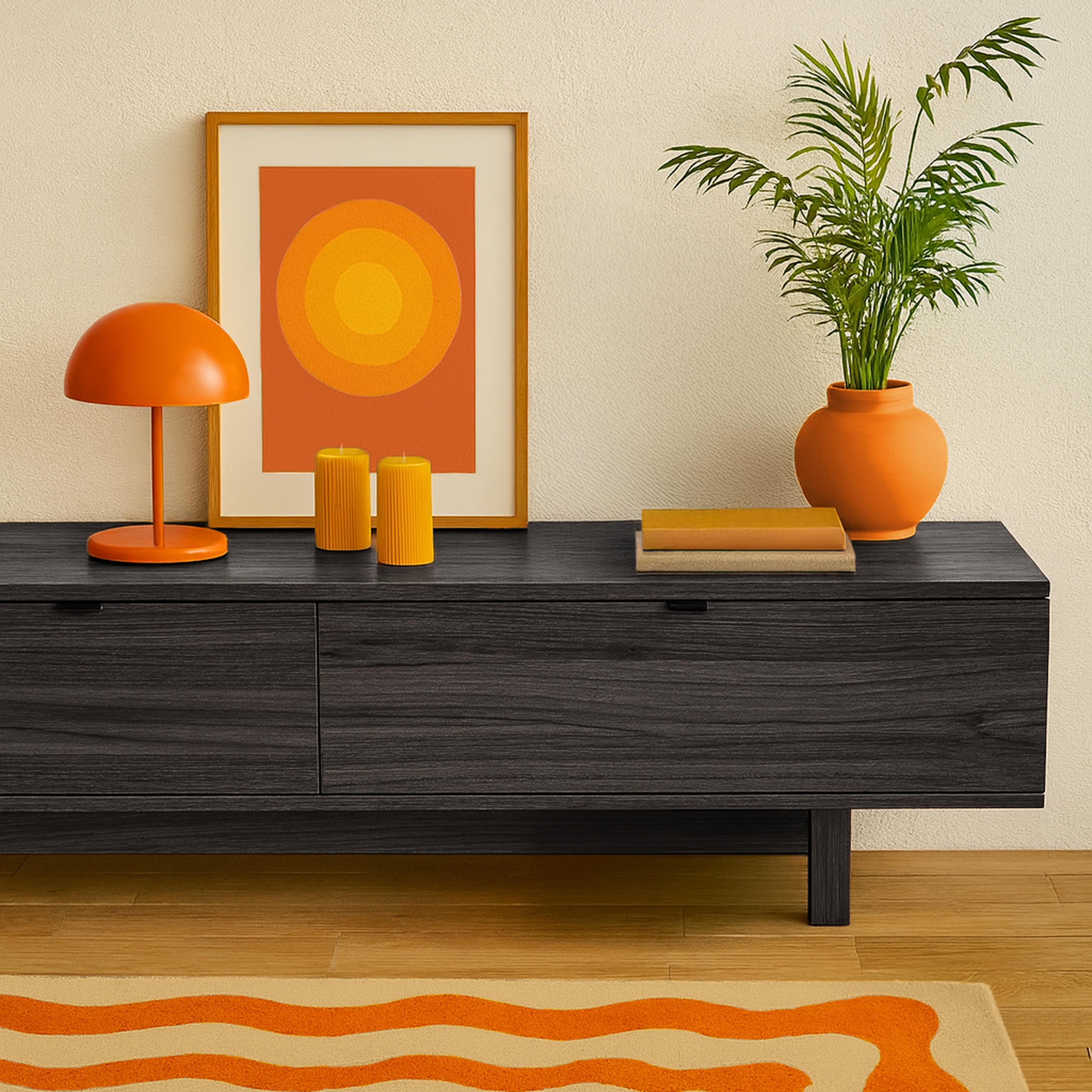 Ein Raum mit dunklem Schrank, orangefarbener Lampe, Kunst, Kerzen, einer Pflanze in einem orangefarbenen Topf und einem Teppich mit orangefarbenen Wellen.