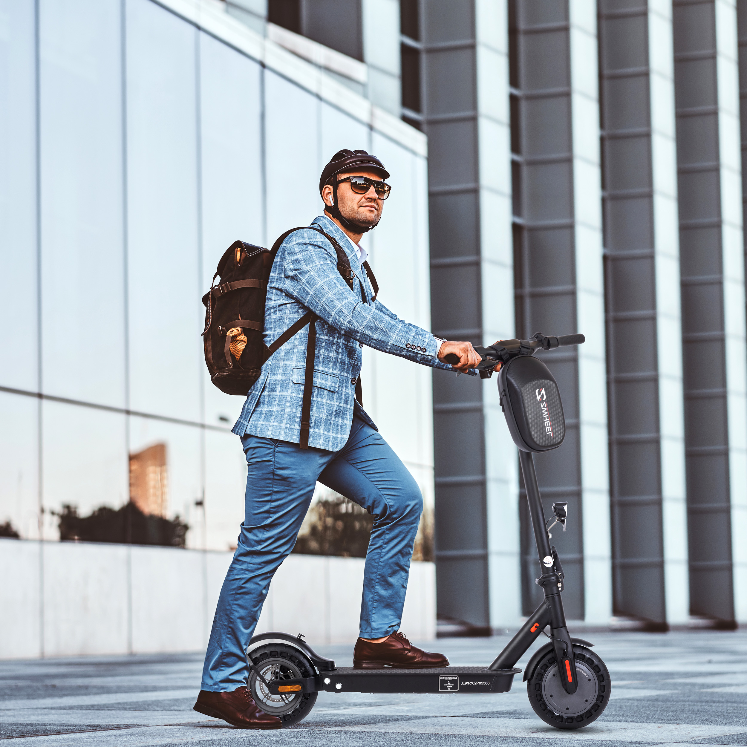 Ein Mann in blauem Anzug fährt einen schwarzen Elektroroller. Er trägt einen Helm und eine Sonnenbrille. Modernes Gebäude im Hintergrund.
