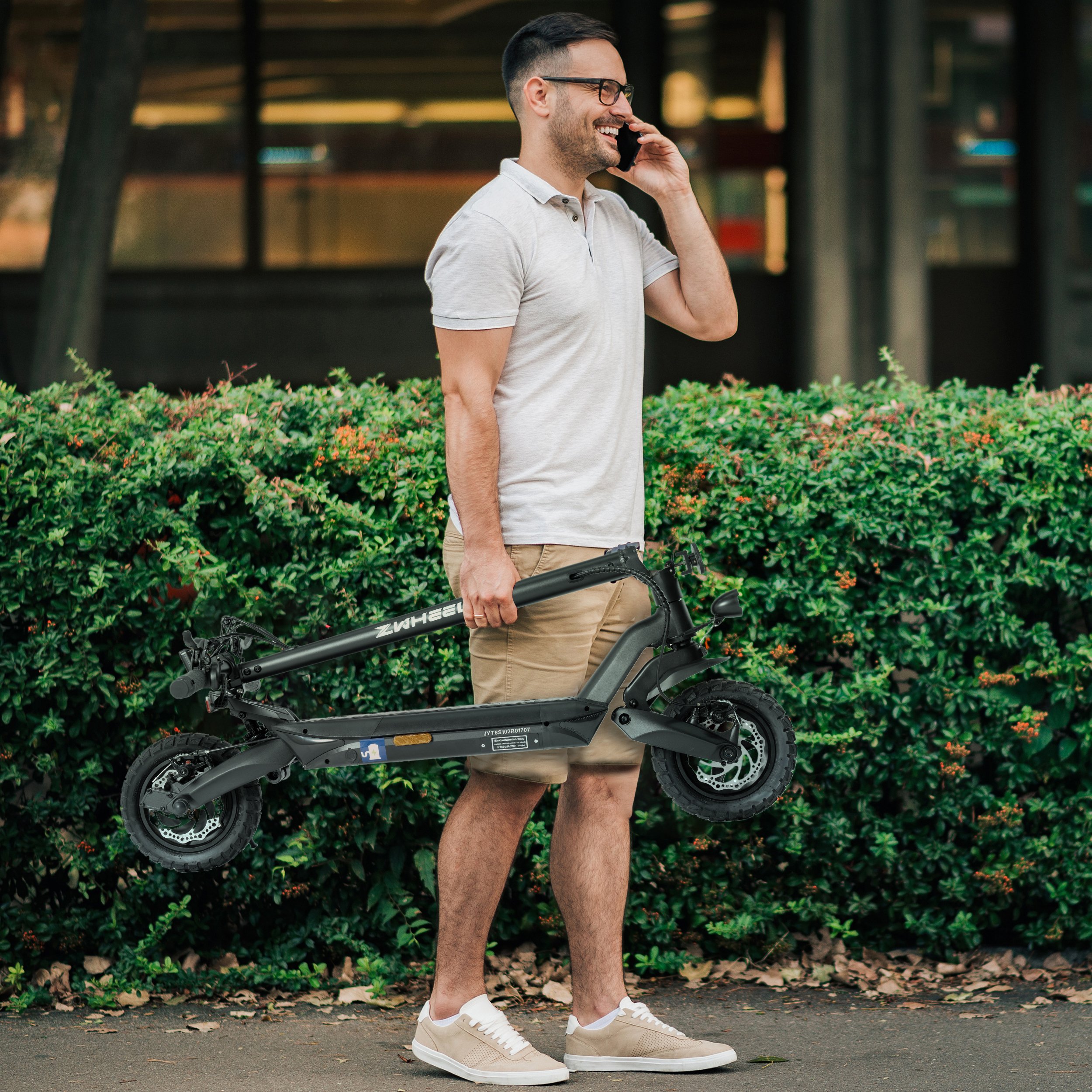 Ein Mann mit Brille telefoniert und hält einen zusammengeklappten schwarzen Roller. Grüne Büsche im Hintergrund.