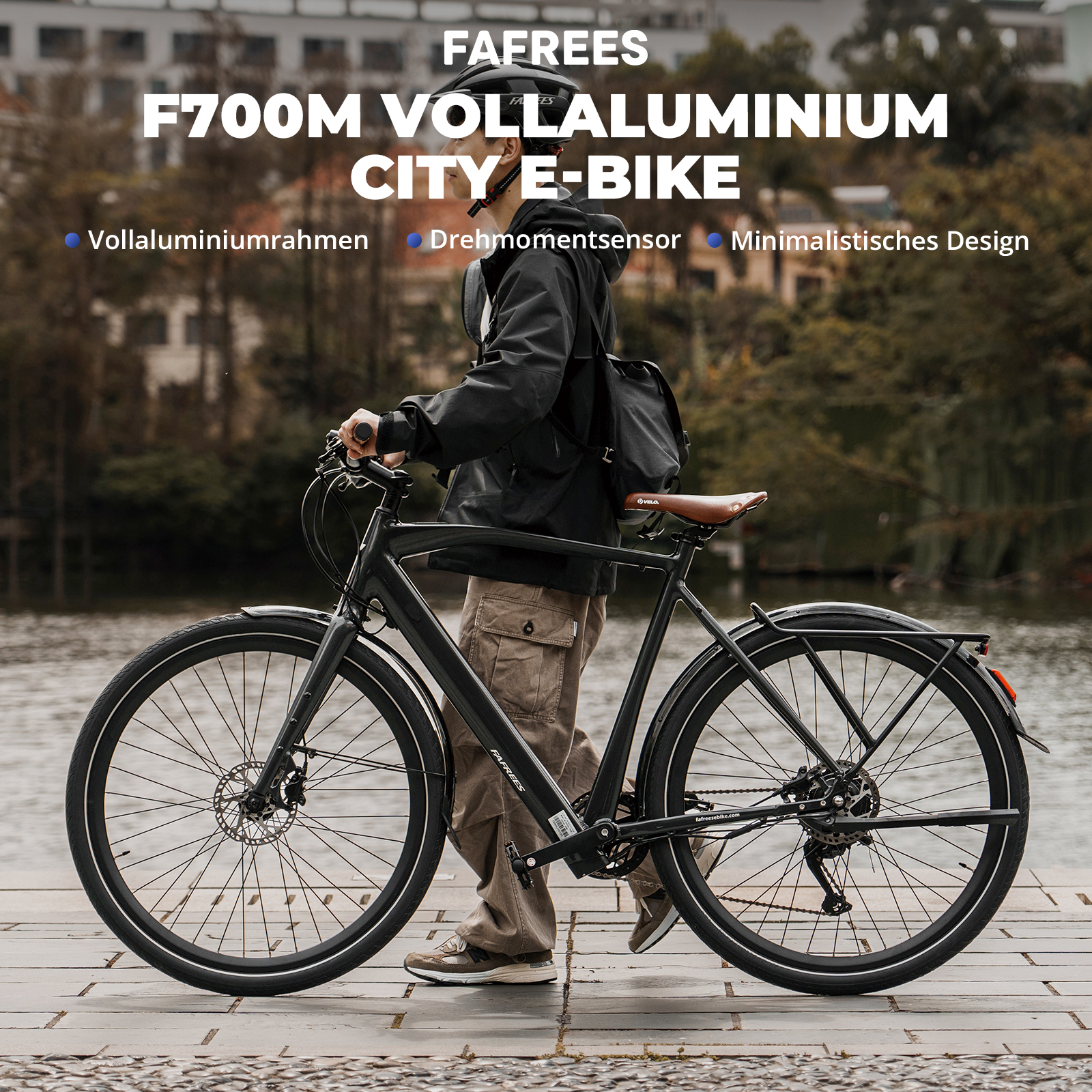 Eine Person in schwarzer Jacke und Helm steht mit einem schwarzen E-Bike. Das Fahrrad steht auf einem Ziegelweg mit Wasser im Hintergrund.