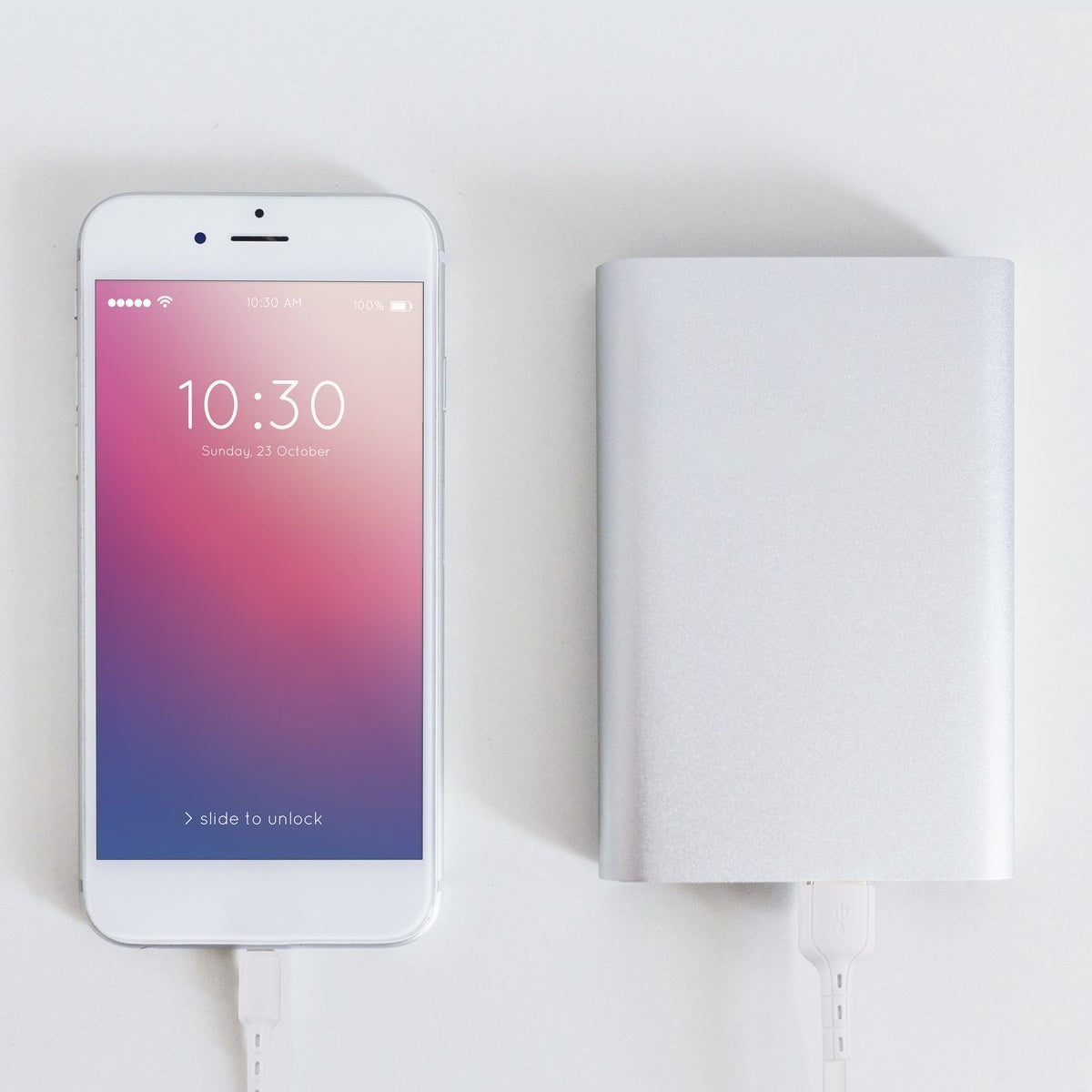 Een witte telefoon die de tijd weergeeft, 10:30, verbonden met een zilveren powerbank met een witte oplaadkabel op een wit oppervlak.