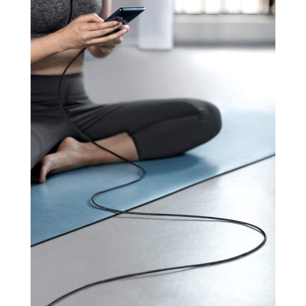 Persoon zit op een yogamat en houdt een telefoon vast. Een zwarte kabel verbindt de telefoon. Lichte scène met neutrale tinten.