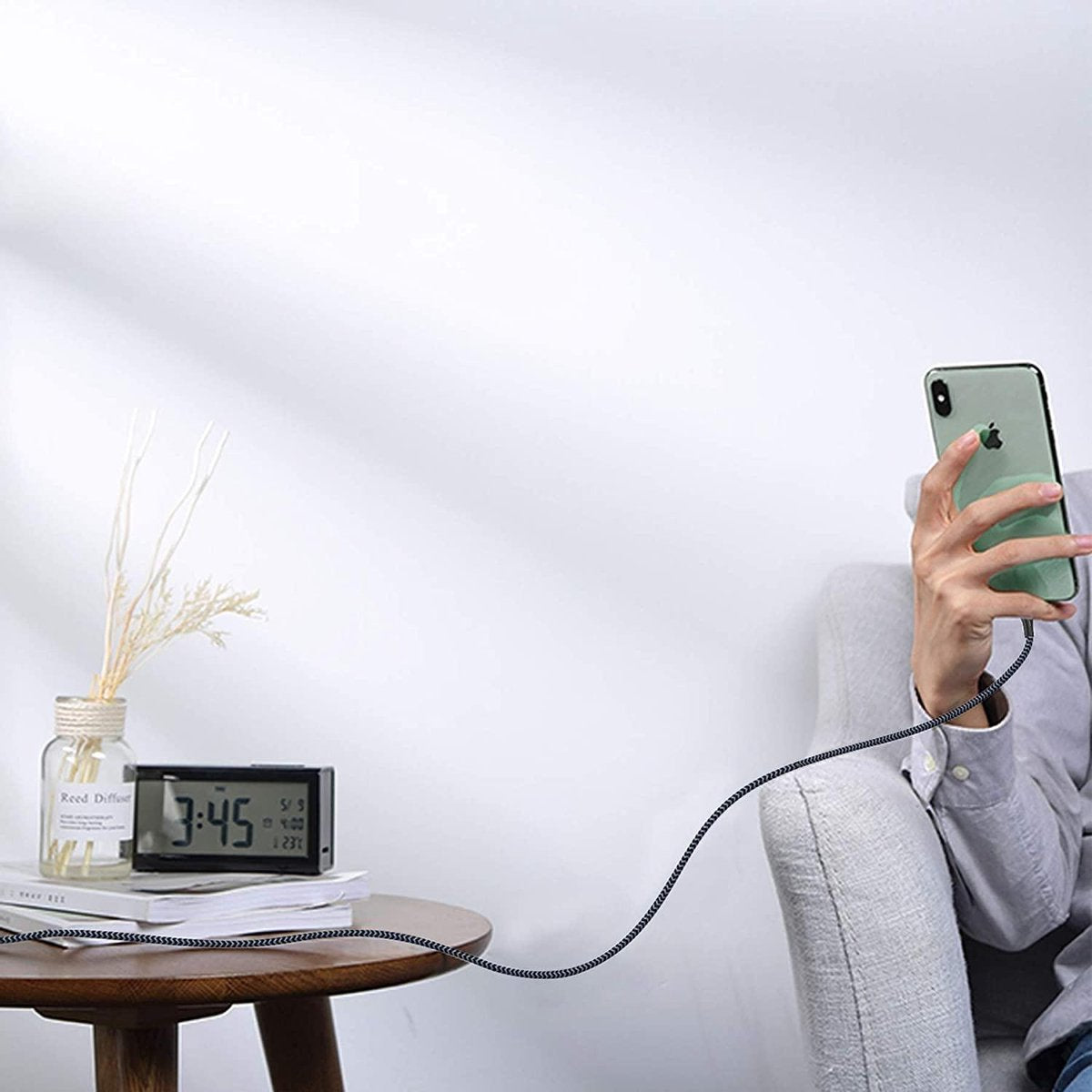 Een persoon houdt een telefoon vast die is aangesloten op een tafel met een klok en een vaas. De achtergrond is een witte muur.