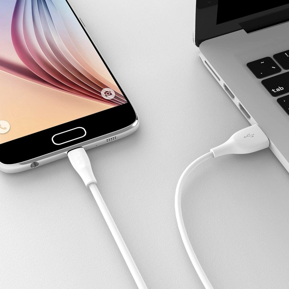 A white USB cable connects a smartphone to a laptop. The phone is on the left and the laptop on the right.
