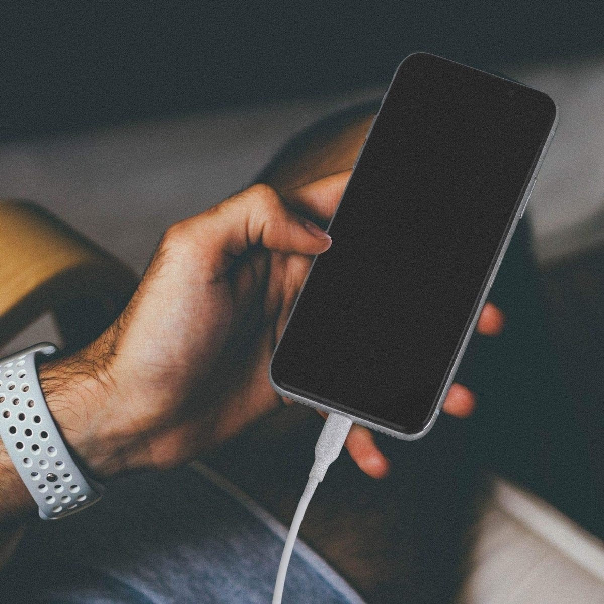Een persoon houdt een smartphone vast, die via een witte kabel wordt opgeladen. Ze draagt een lichtgrijs horloge. De achtergrond is wazig.