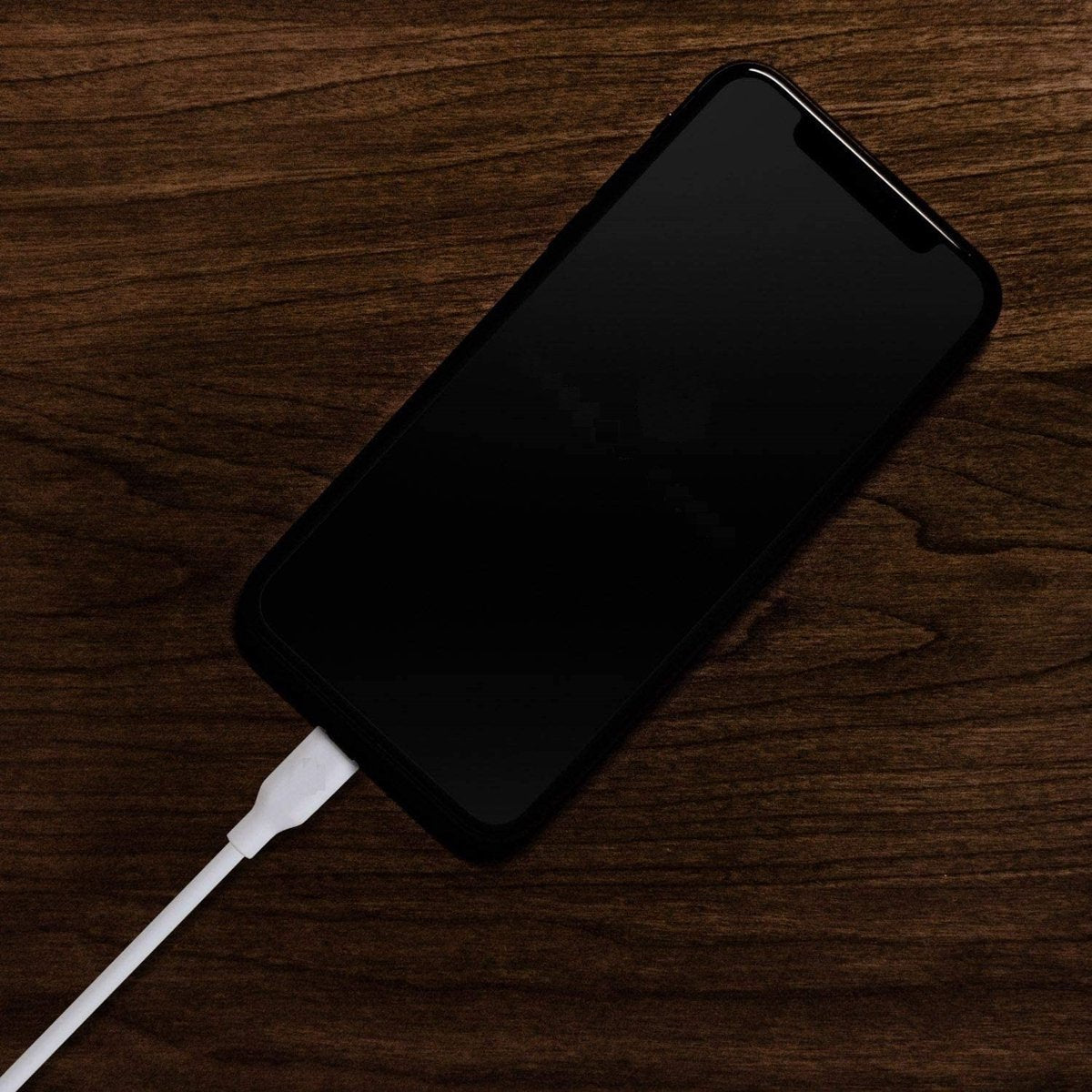 Een zwarte smartphone ligt op een donker houten oppervlak. Een witte oplaadkabel is aangesloten op de telefoon. De achtergrond is van hout.