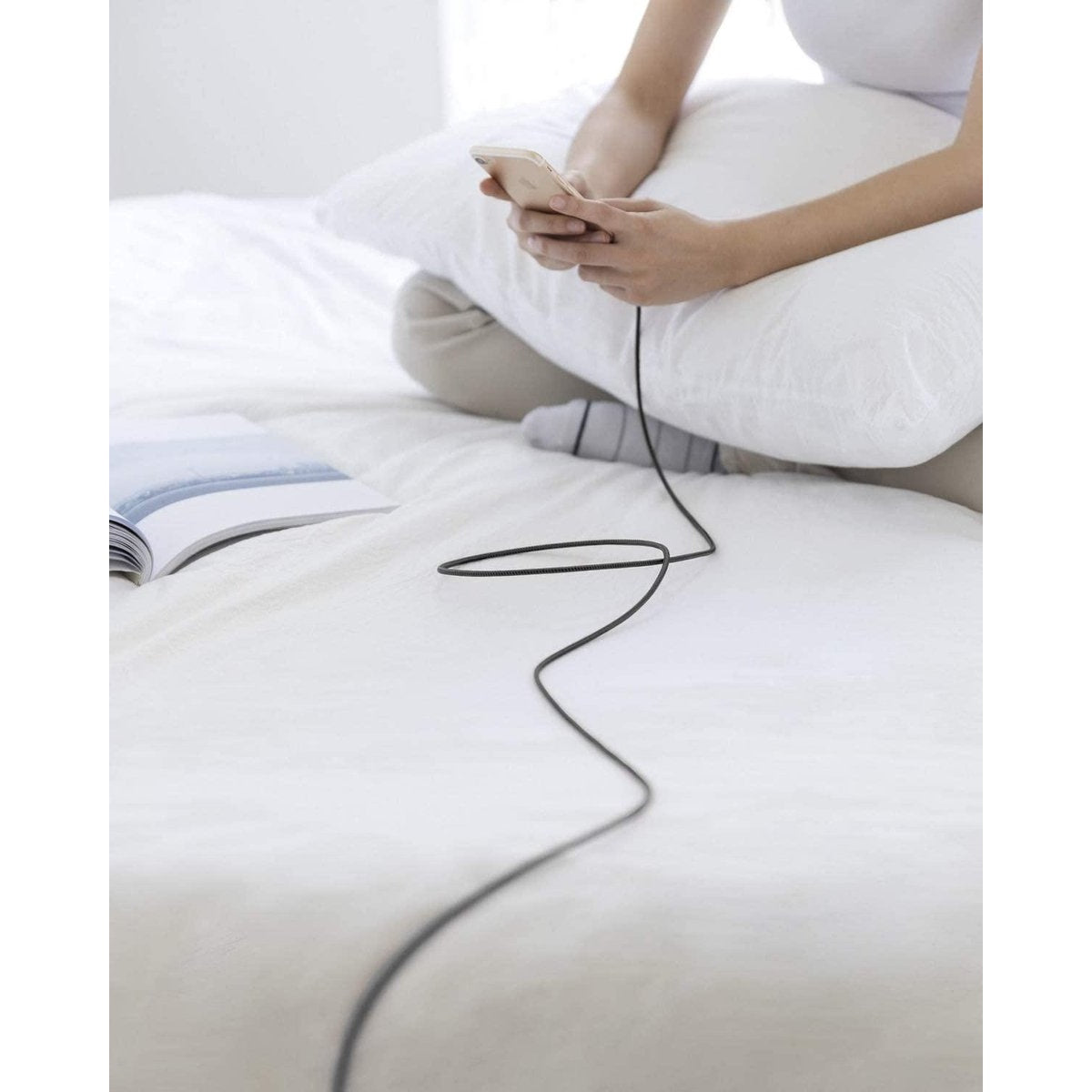 A person sitting on a bed with a phone connected to a cable. A book lies open. White bedding and a pillow are visible.