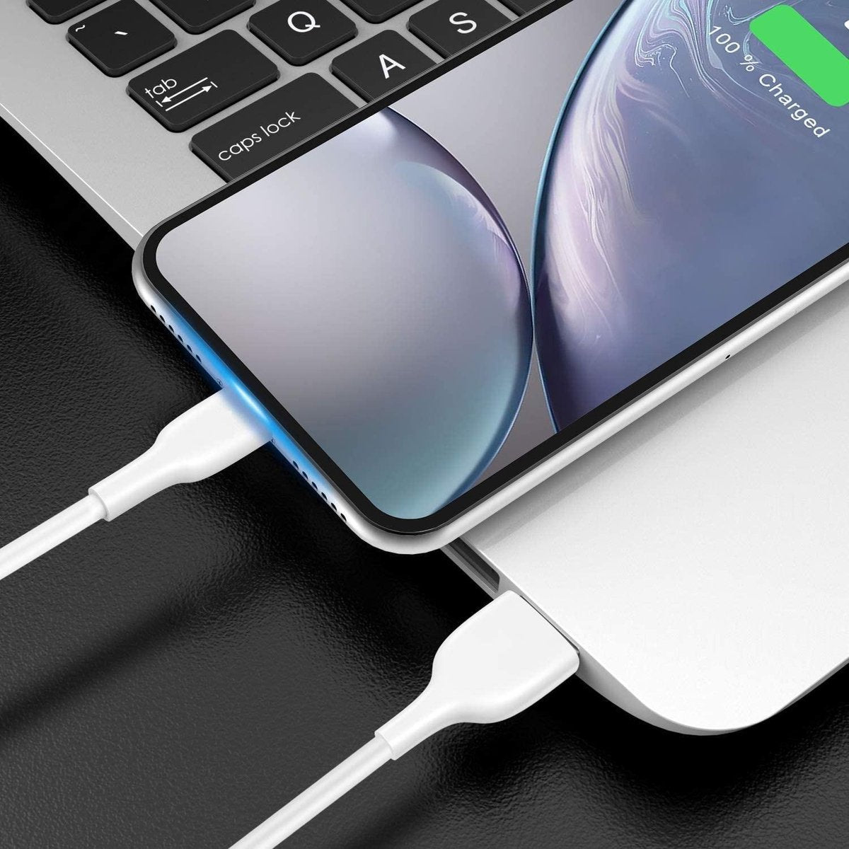 A smartphone charging on a white surface, connected by a white cable. The phone displays 100% charged on the screen. A laptop keyboard is visible.