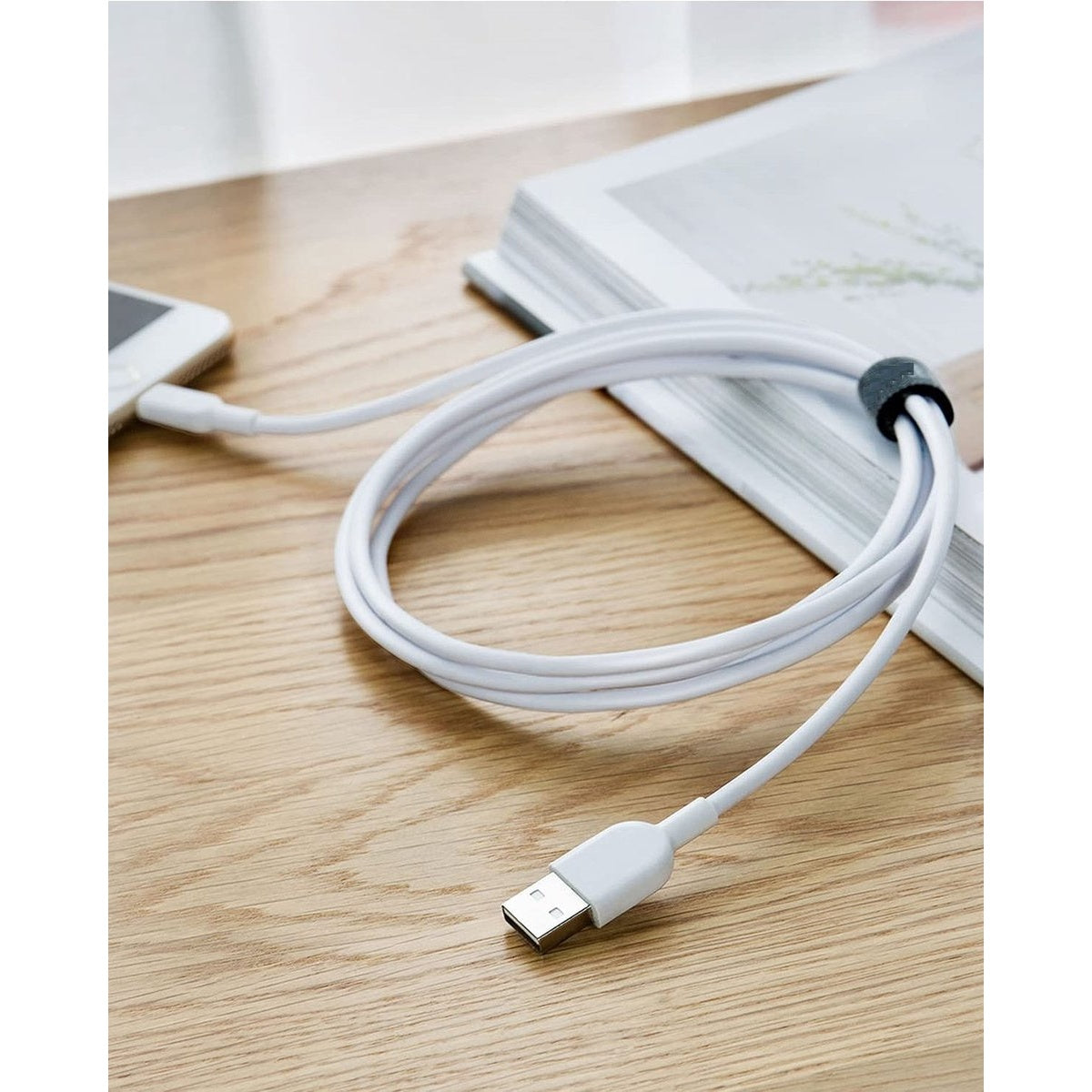 Witte USB-kabel aangesloten op een telefoon en gewikkeld rond een boek op een houten tafel. De kabel wordt vastgehouden door een zwarte band.