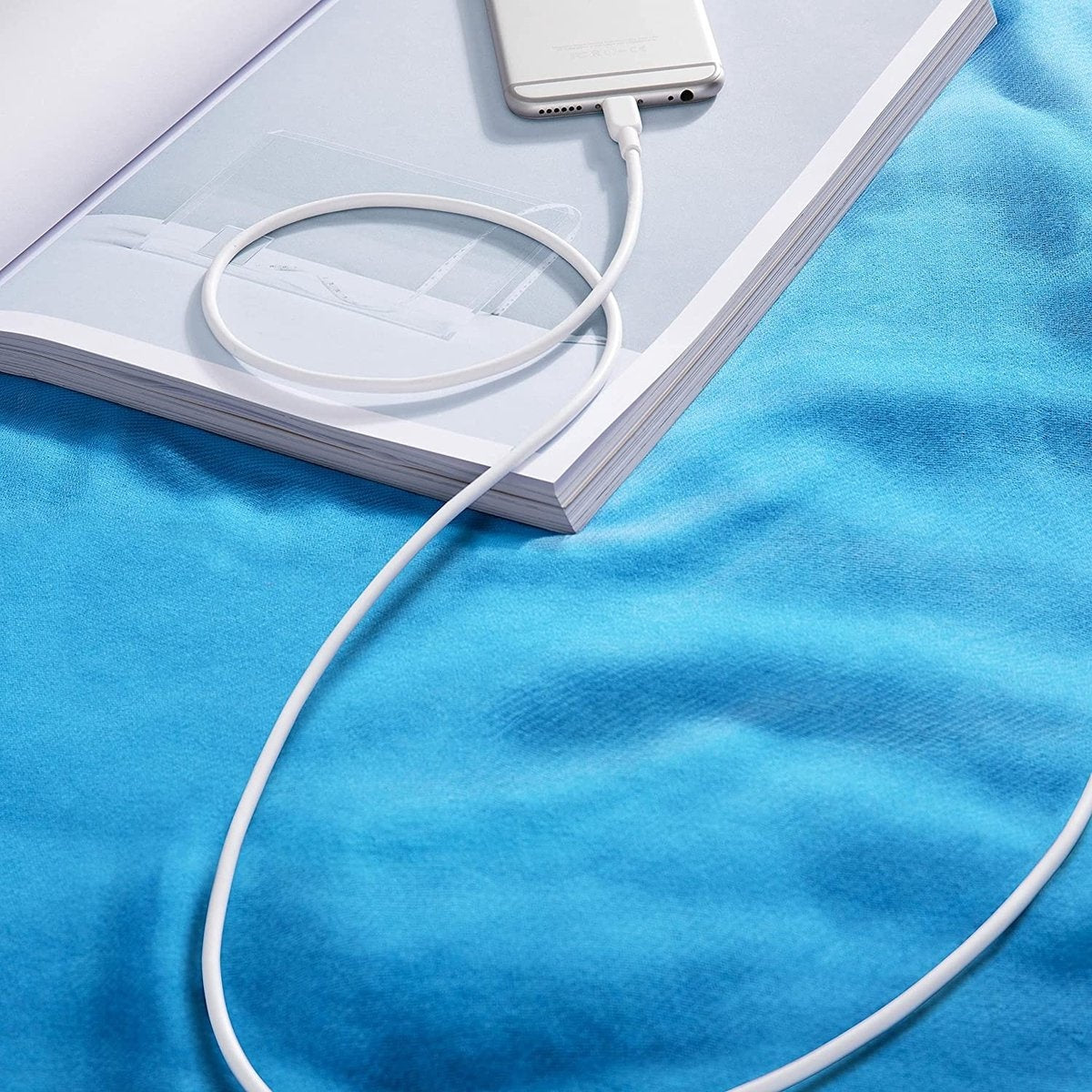 Een witte telefoon laadt op een boek met een witte kabel. Het boek ligt op een blauwe stoffen achtergrond.