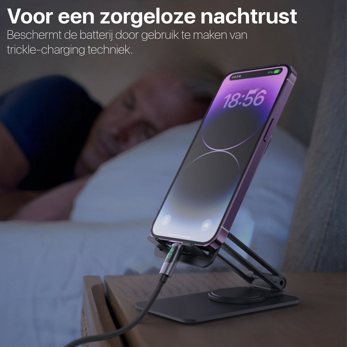 Een telefoon die oplaadt op een standaard naast een bed. Een persoon slaapt op de achtergrond. De telefoon toont de tijd 18:56.