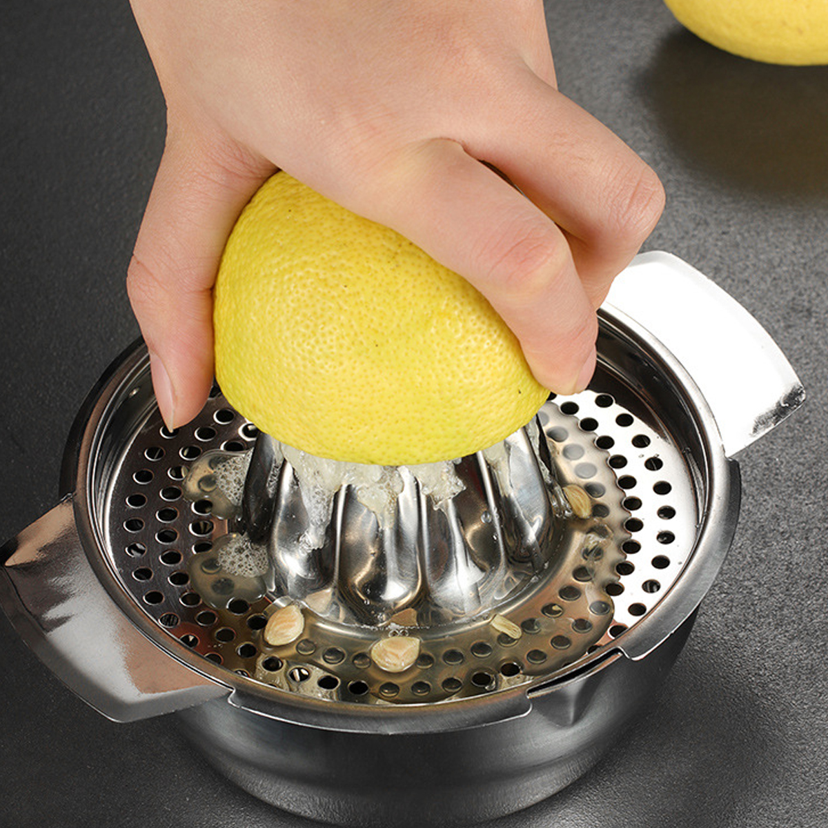 Eine Hand presst eine Zitrone über einer Saftpresse aus. Die Zitrone, gelb, ist halbiert. Zitronensaft und Kerne sind in der Saftpresse zu sehen.