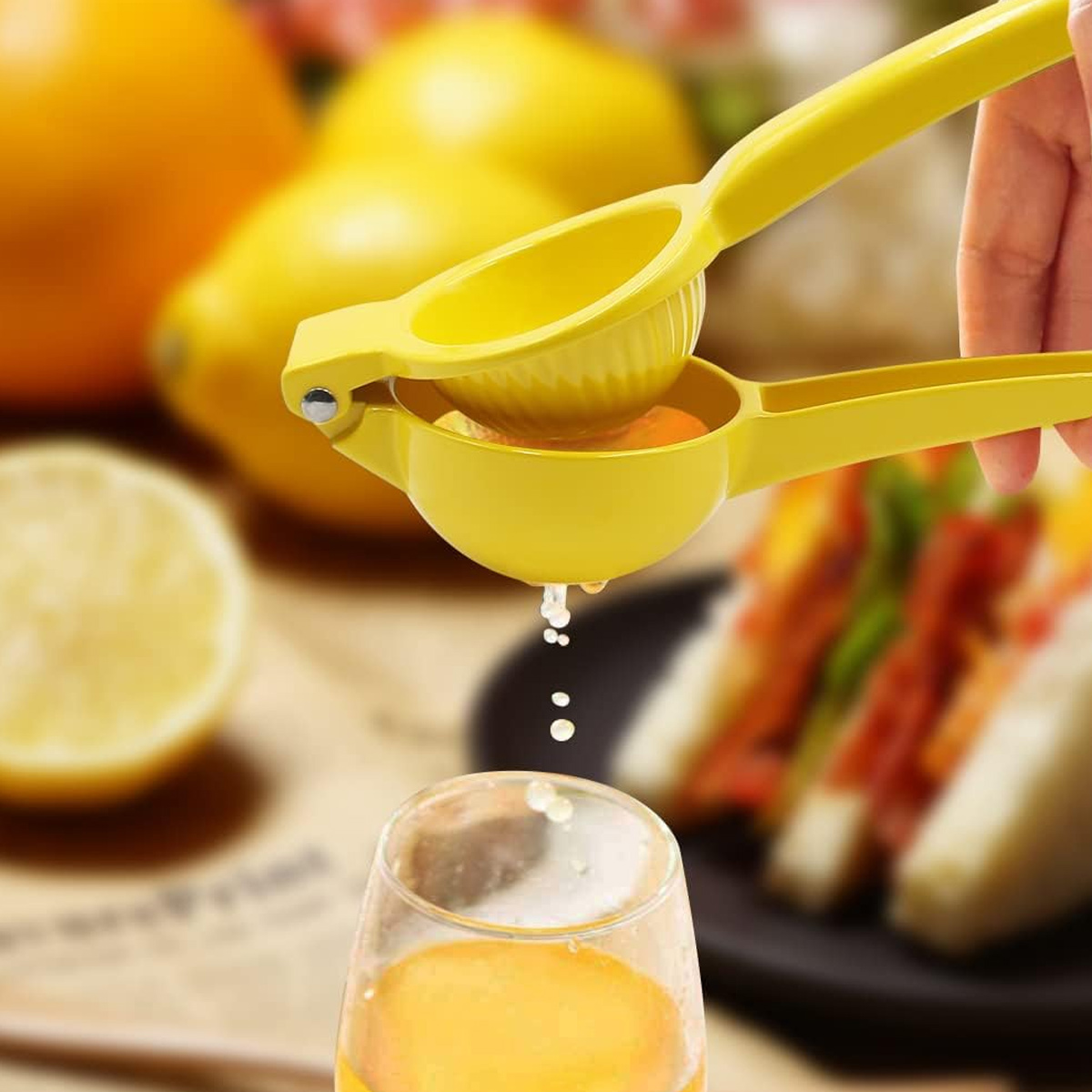 Eine Hand presst mit einem gelben Entsafter eine Zitrone, Saft tropft in ein Glas. Zitronen und Sandwiches sind im Hintergrund.