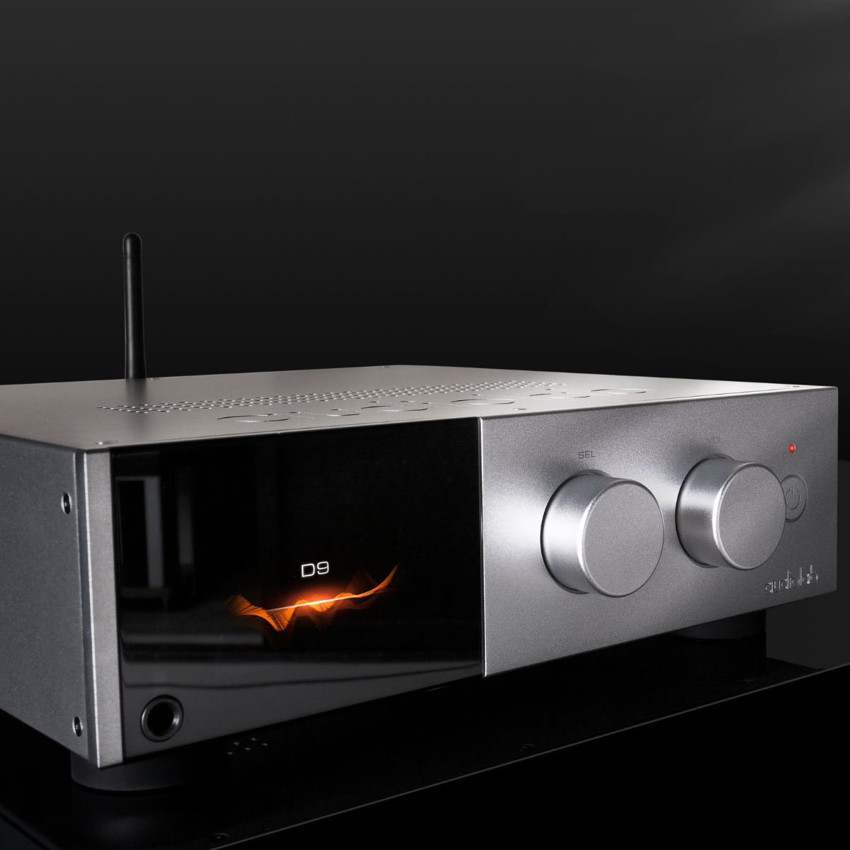 Een zilveren audioversterker met knoppen en een display met oranje graphics. Het heeft een zwarte achtergrond en een kleine antenne.