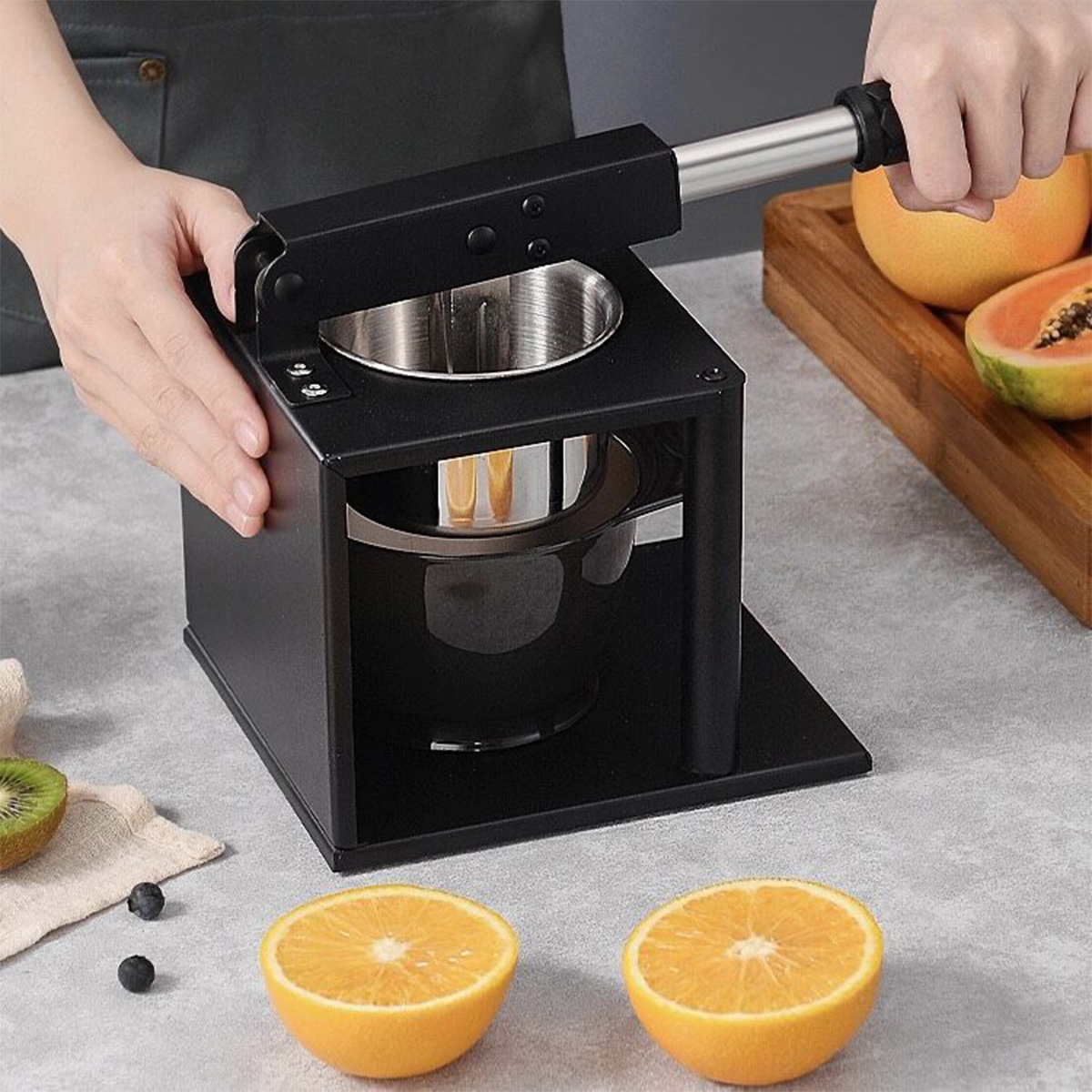 Eine Person benutzt eine schwarze Saftpresse. In der Nähe befinden sich halbierte Orangen, Kiwis und Grapefruits. Ein Holztablett enthält mehr Obst.