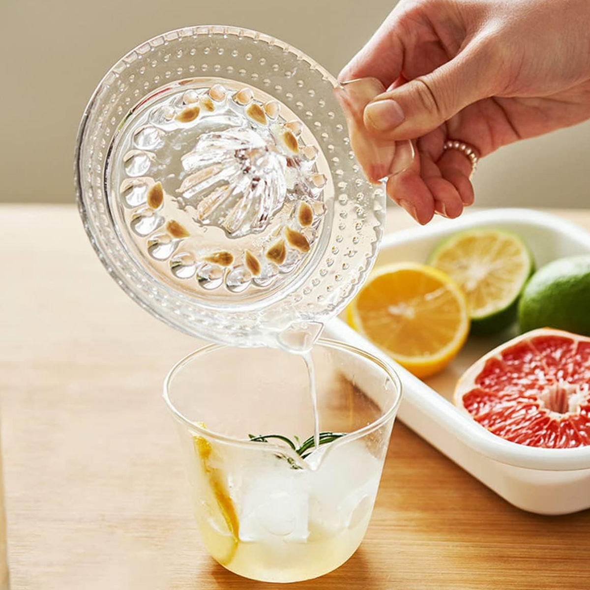Eine Hand presst Saft in ein Glas mit Eis und Zitrone. Zitrusfrüchte liegen auf einem weißen Tablett. Glasentsafter im Vordergrund.