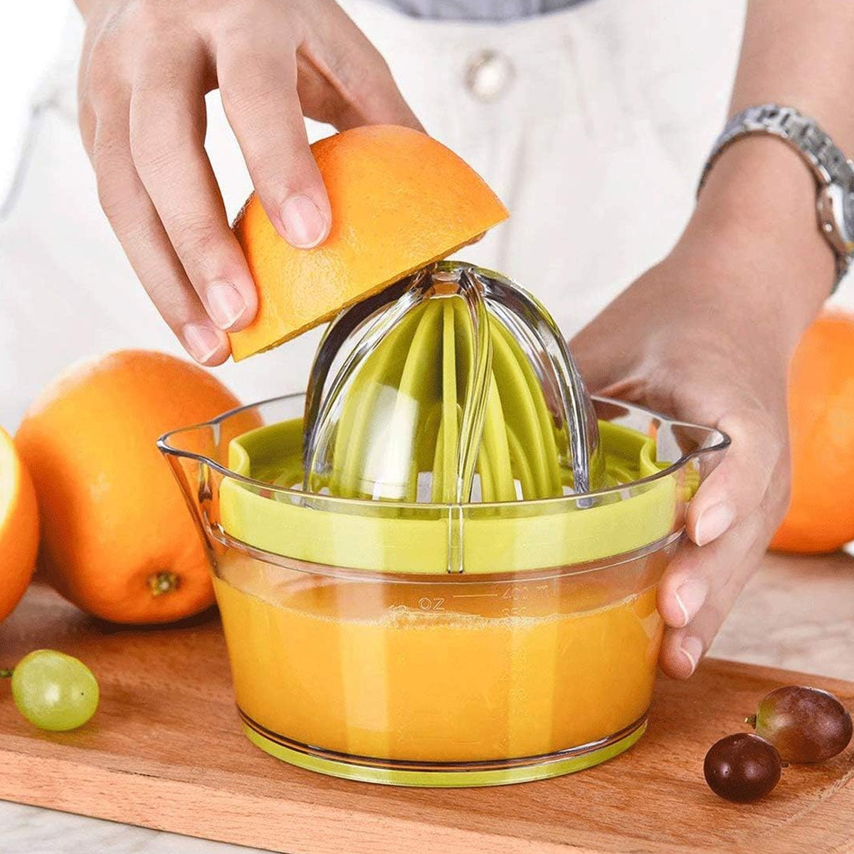 Hände, die eine Orangenhälfte über einer Saftpresse ausquetschen. Der Entsafter ist mit Orangensaft gefüllt. Trauben und Orangen liegen auf dem Holzbrett.
