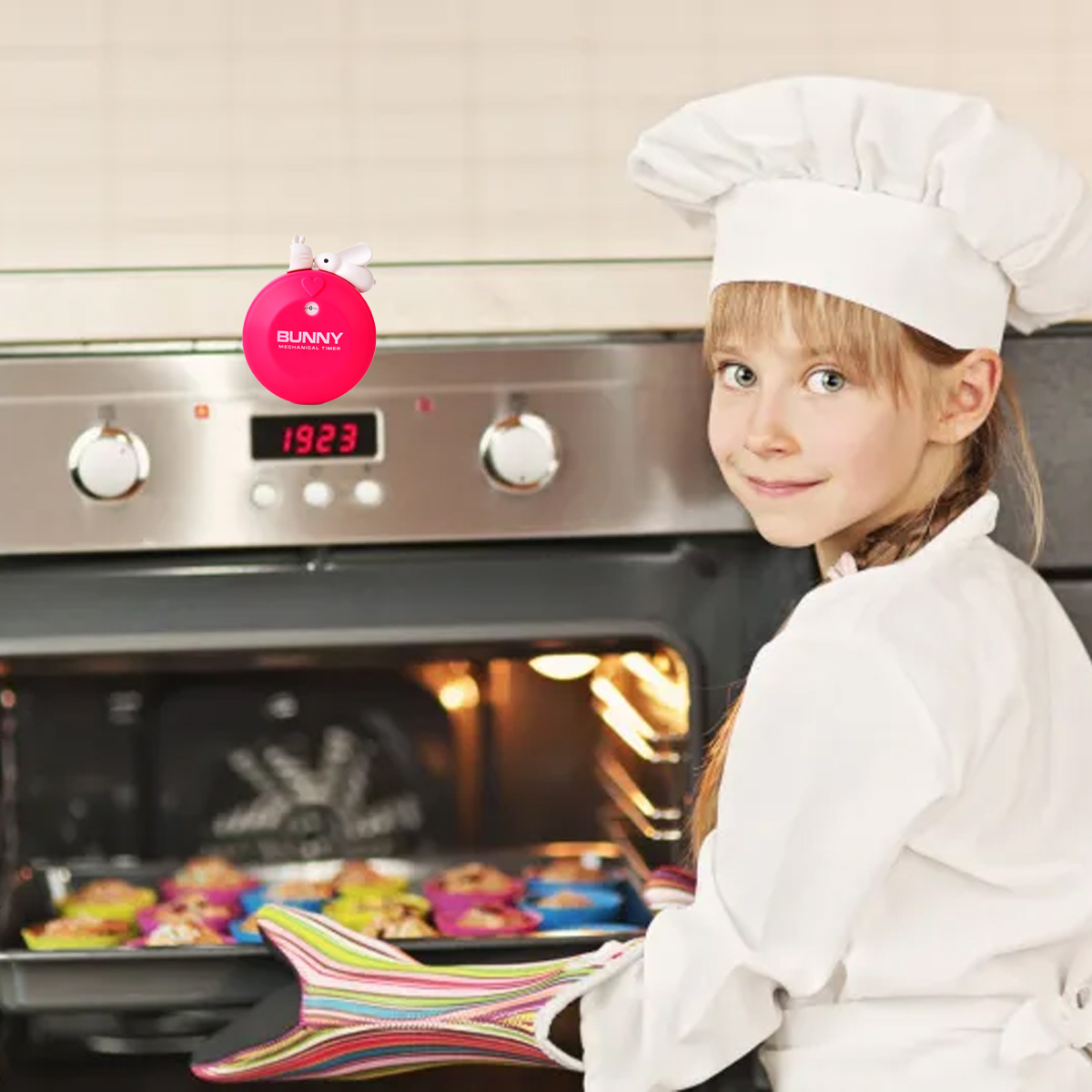 Ein junges Mädchen mit Kochmütze und Schürze lächelt. Sie hält Ofenhandschuhe, im Hintergrund backen Muffins im Ofen.