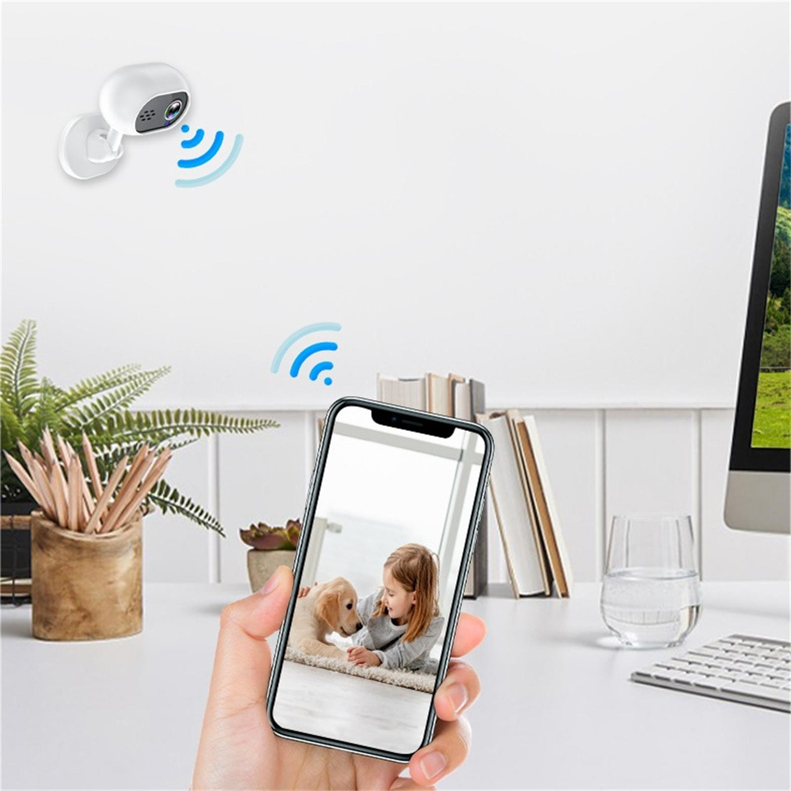 Eine Hand hält ein Telefon, das ein Mädchen mit einem Hund zeigt. Eine Überwachungskamera und ein WLAN-Signal sind sichtbar. Andere Objekte sind auf dem weißen Schreibtisch.
