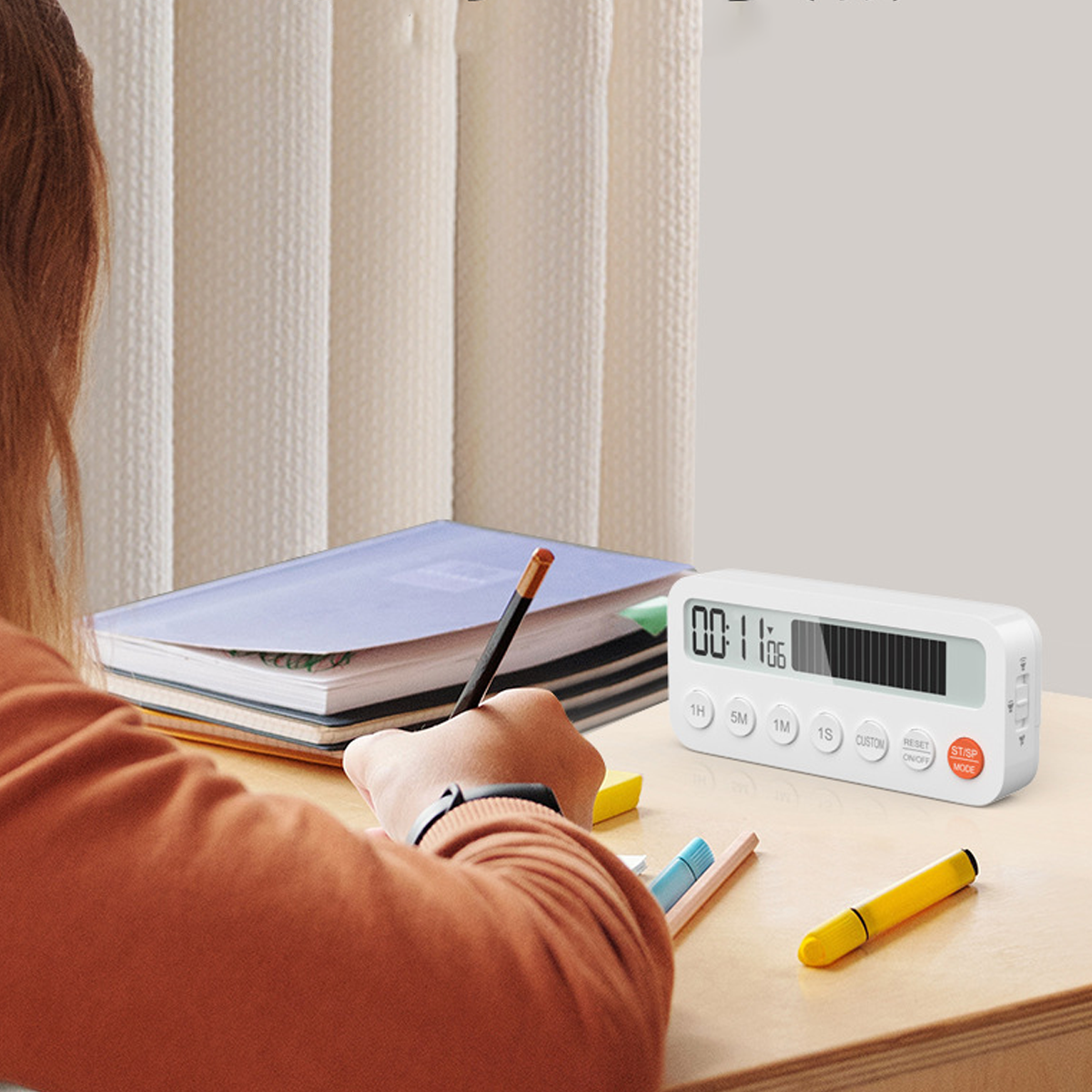 Eine Person schreibt mit einem Bleistift auf einem Schreibtisch. Ein Timer zeigt die Zeit an. Bücher, Stifte und ein gelber Textmarker liegen auf dem Schreibtisch.