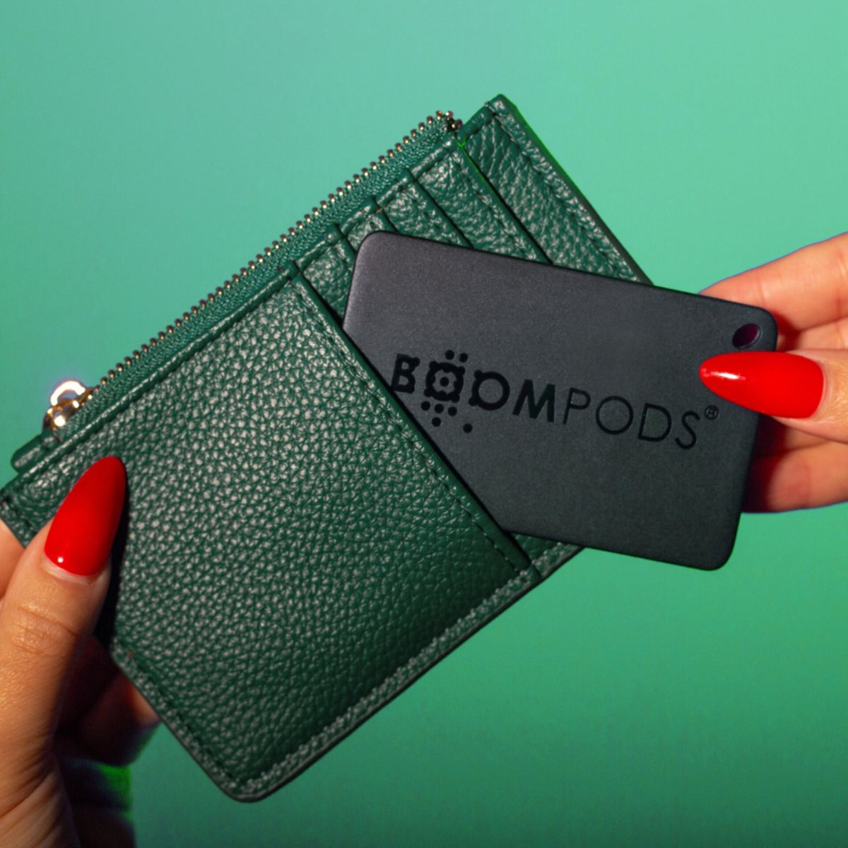 Mani che tengono un portafoglio verde e una carta nera con scritto BOOMPODS. Smalto rosso. Sfondo verde.