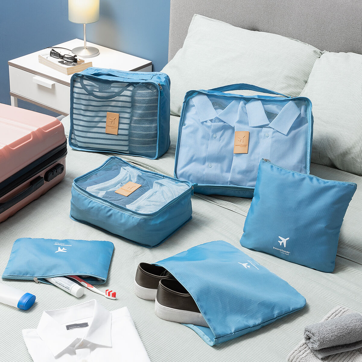 Blaue Packwürfel und -taschen mit Kleidung, Schuhen und Toilettenartikeln auf einem Bett. Ein rosa Koffer ist links.