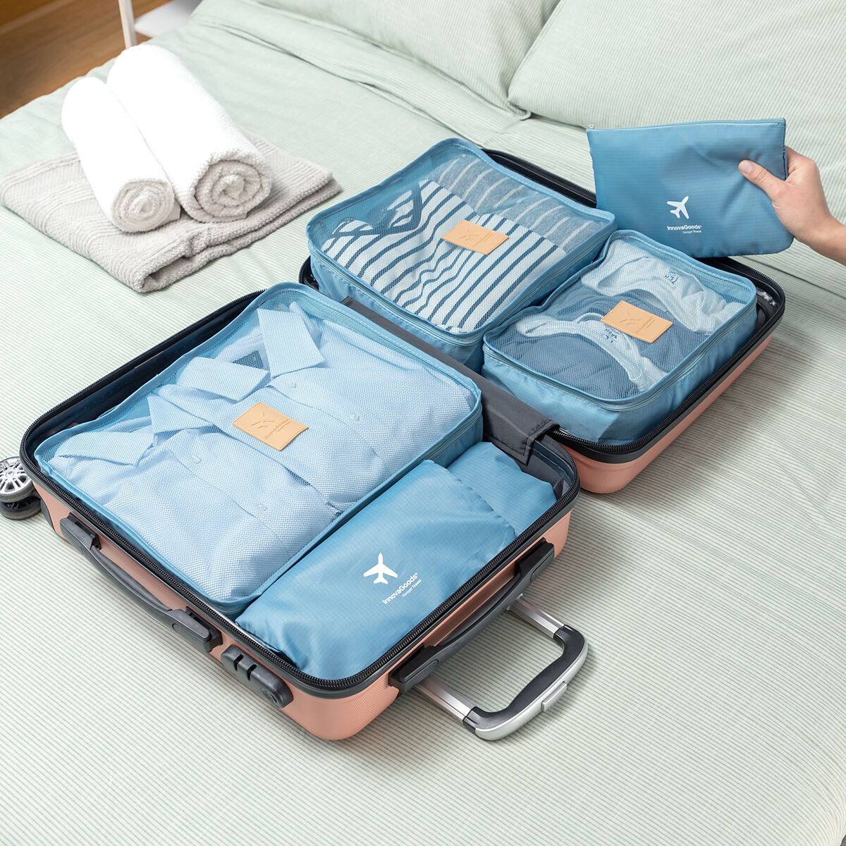 Offener Koffer gefüllt mit Packwürfeln und Kleidung. Eine Hand hält einen kleinen blauen Beutel. Handtücher liegen im Hintergrund auf dem Bett.