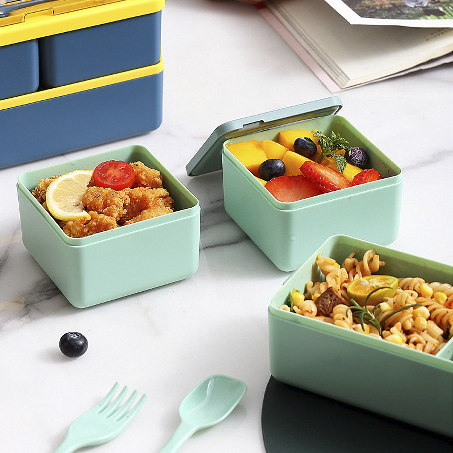 Cajas de almuerzo con comida sobre una superficie de mármol. Las cajas contienen pollo frito, frutas y pasta. Un tenedor y una cuchara están cerca.