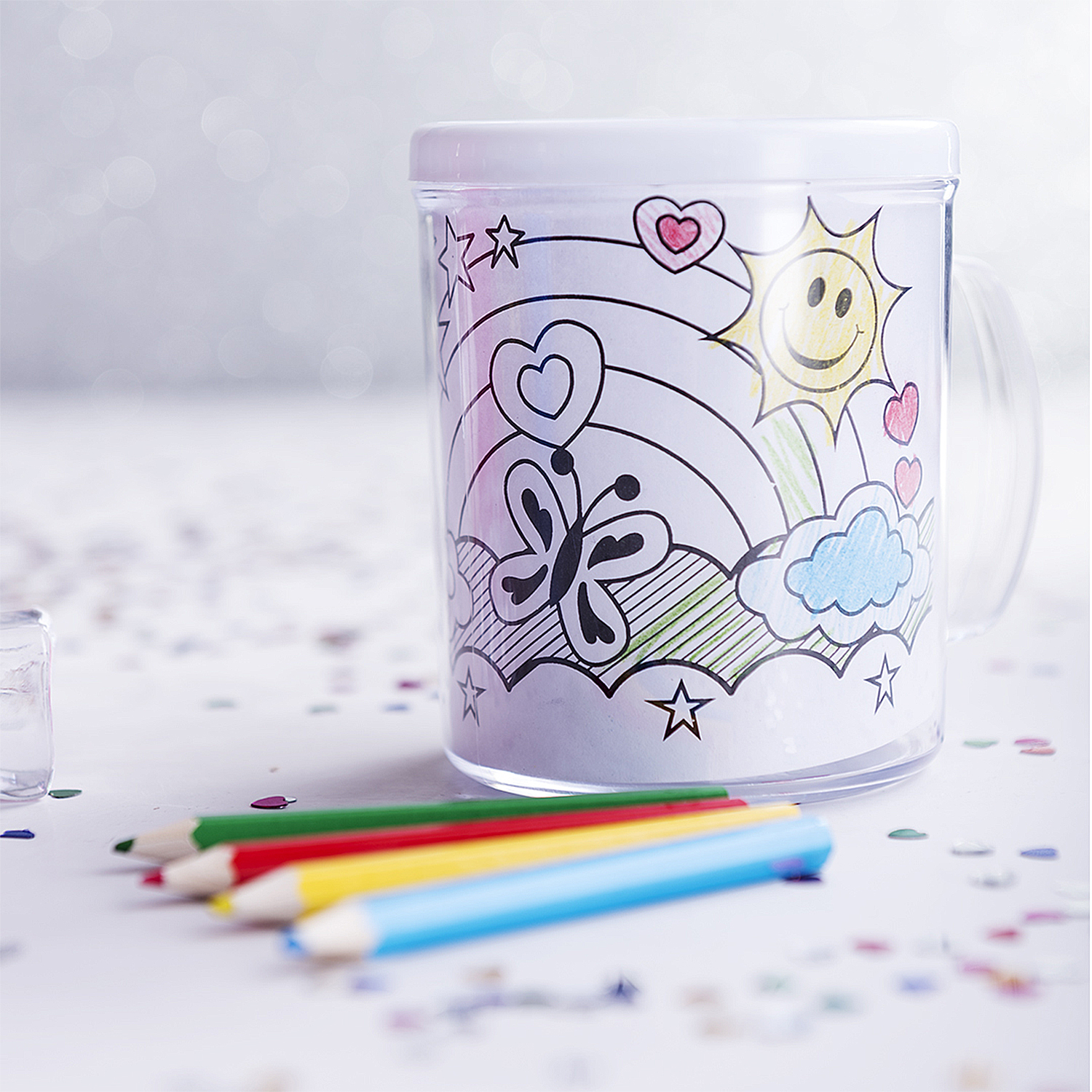 Una tazza trasparente con un disegno di sole, arcobaleno e farfalla. Matite colorate sono davanti alla tazza.