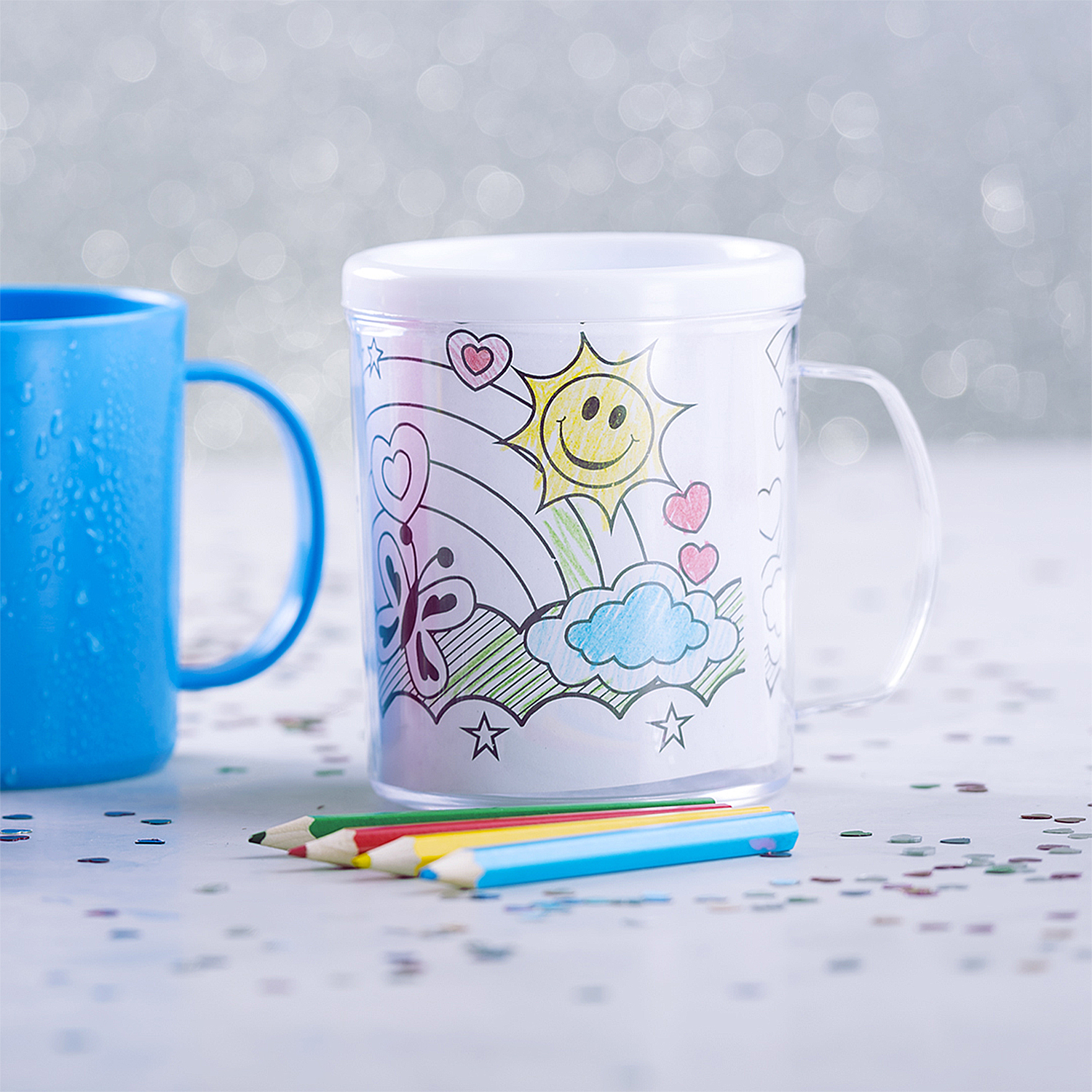 Una tazza blu e una tazza trasparente con un disegno di sole, cuori e farfalla. Matite colorate davanti.