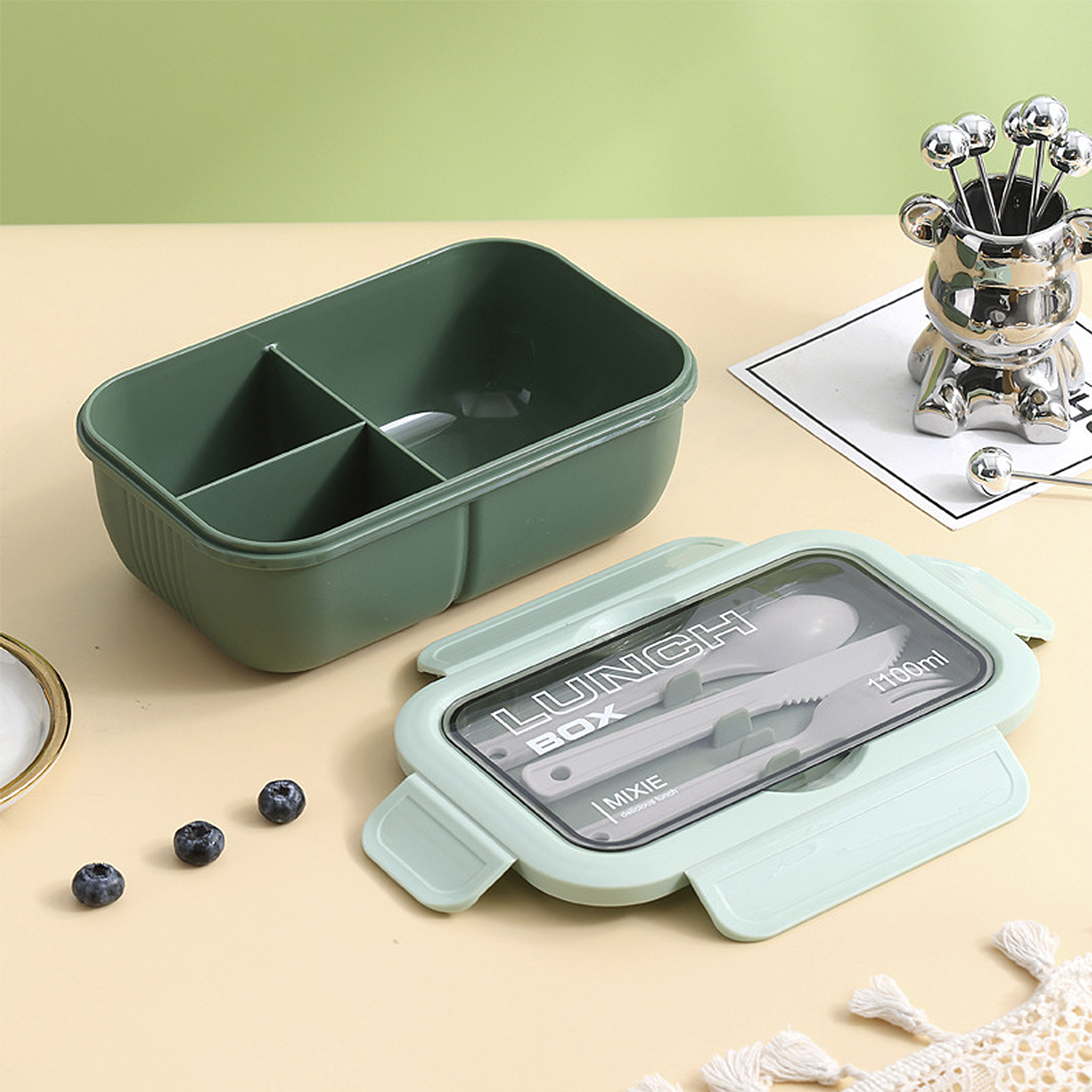 Una fiambrera verde con cubiertos, arándanos y un soporte en forma de oso con pequeñas bolas plateadas sobre una superficie beige.