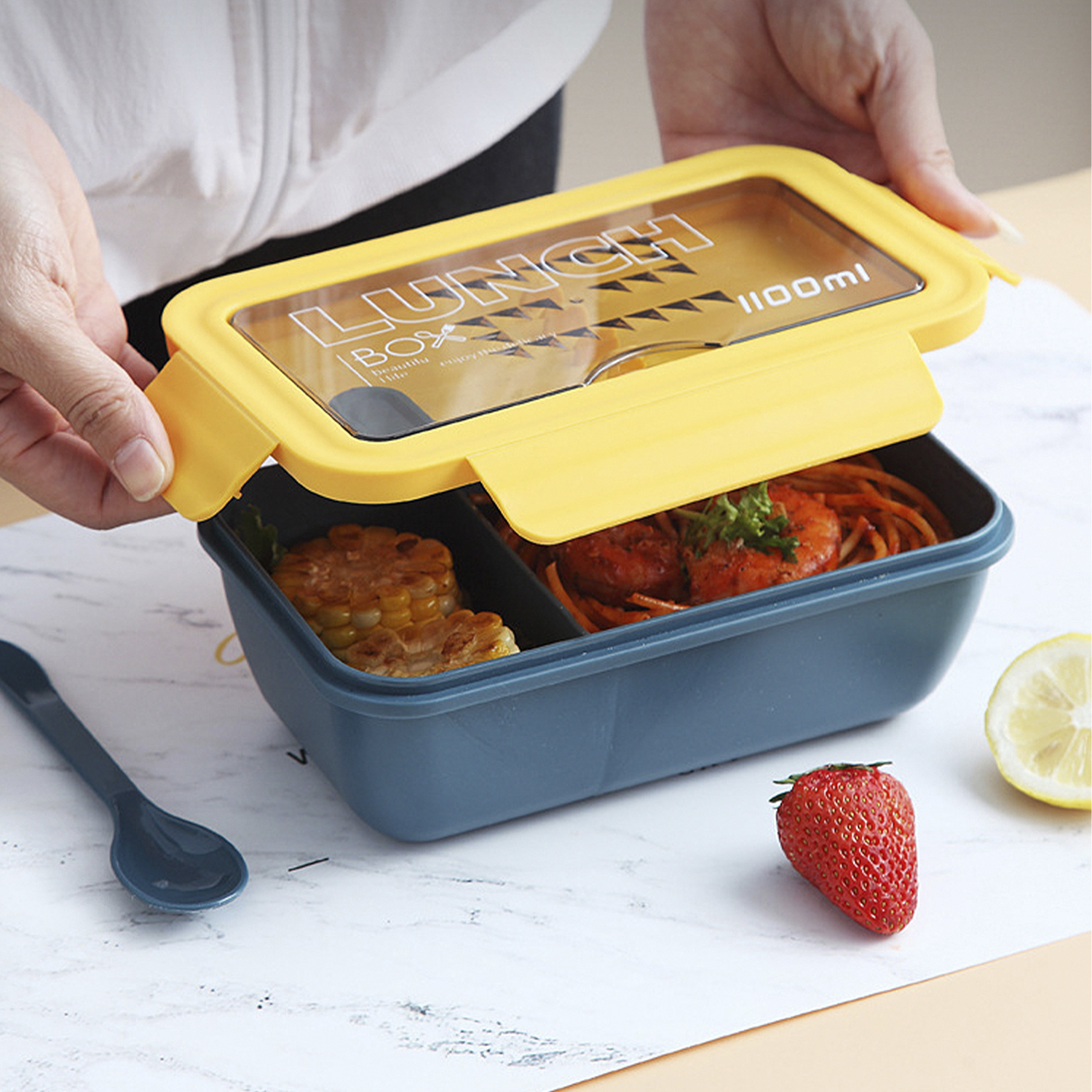 Manos abren una fiambrera con comida: maíz, pasta, camarones y fresa. Una cuchara y limón están junto a la caja sobre una superficie blanca.