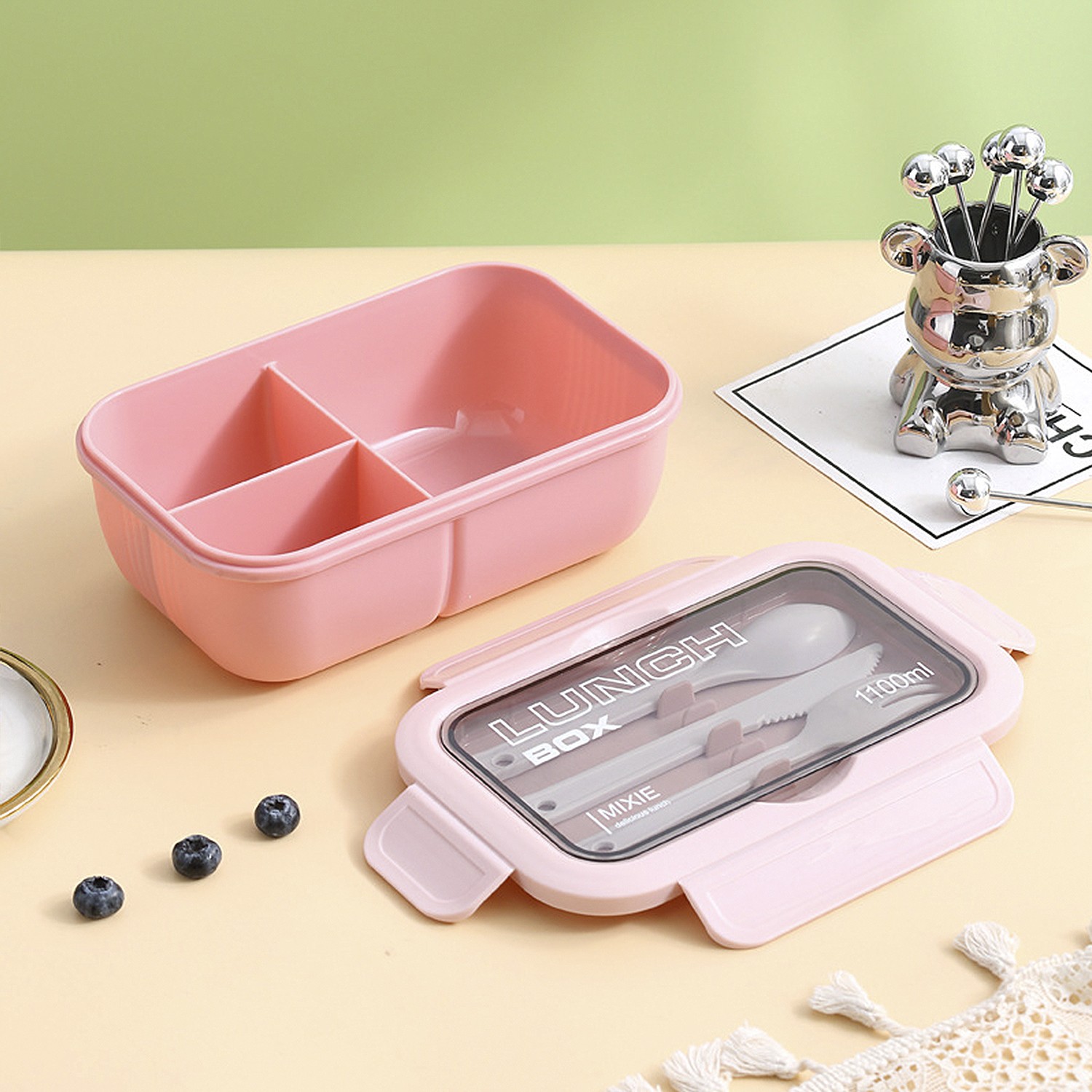 Un portapranzo rosa con posate è aperto. Sono presenti anche mirtilli e un supporto a forma di orso con spiedini d'argento.
