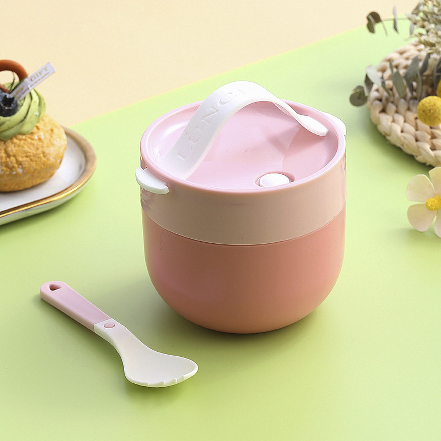 Un portapranzo rosa e bianco con manico e cucchiaio su una superficie verde. Torta a sinistra. Fiori sullo sfondo.