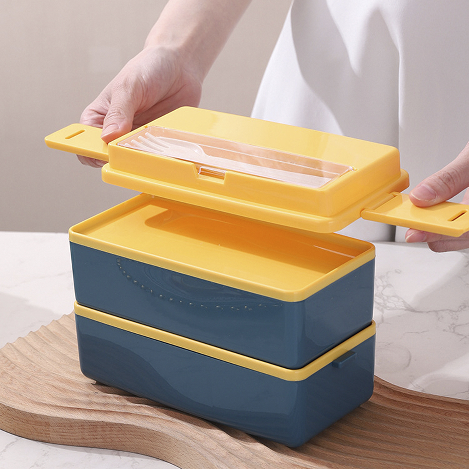Manos abriendo una fiambrera de dos niveles. La caja es azul y amarilla, con una sección de tapa transparente que contiene un tenedor.