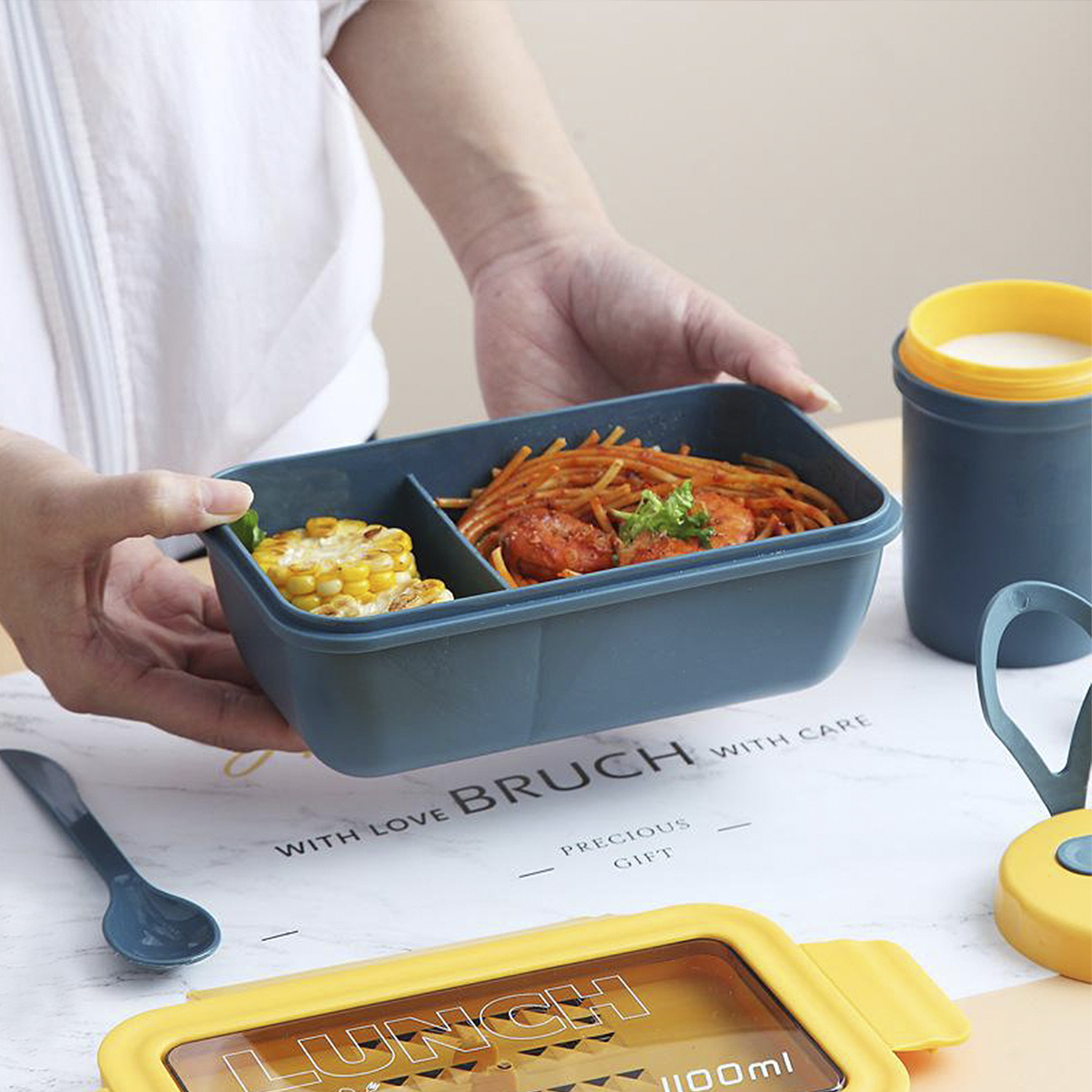 Una persona sostiene una fiambrera dividida con pasta y maíz. Cerca hay un recipiente para bebidas, una cuchara y otra fiambrera. El fondo es una mesa.