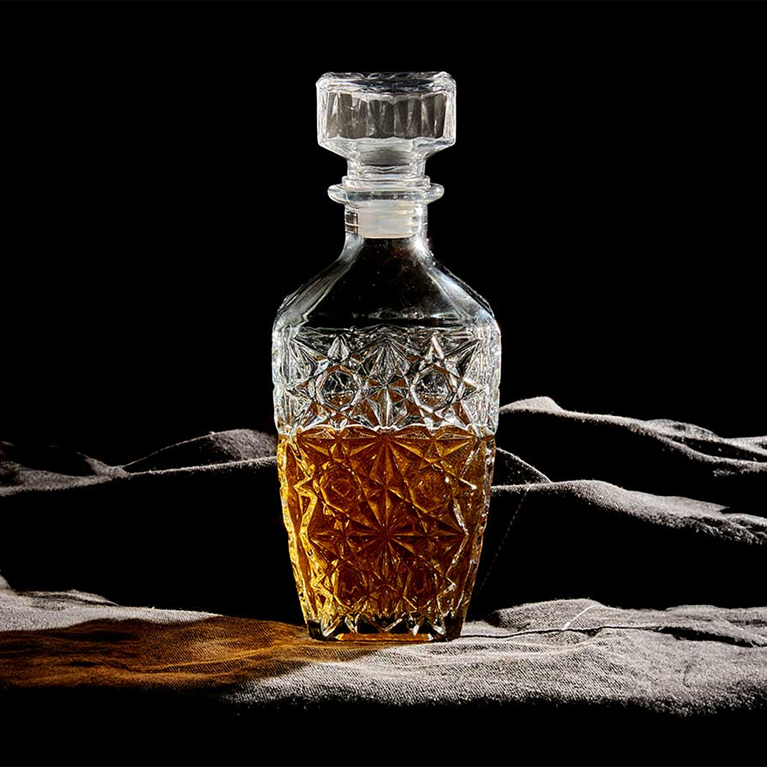 Una caraffa di cristallo con liquido ambrato su un tessuto scuro. La caraffa è trasparente, con un design strutturato e un tappo.