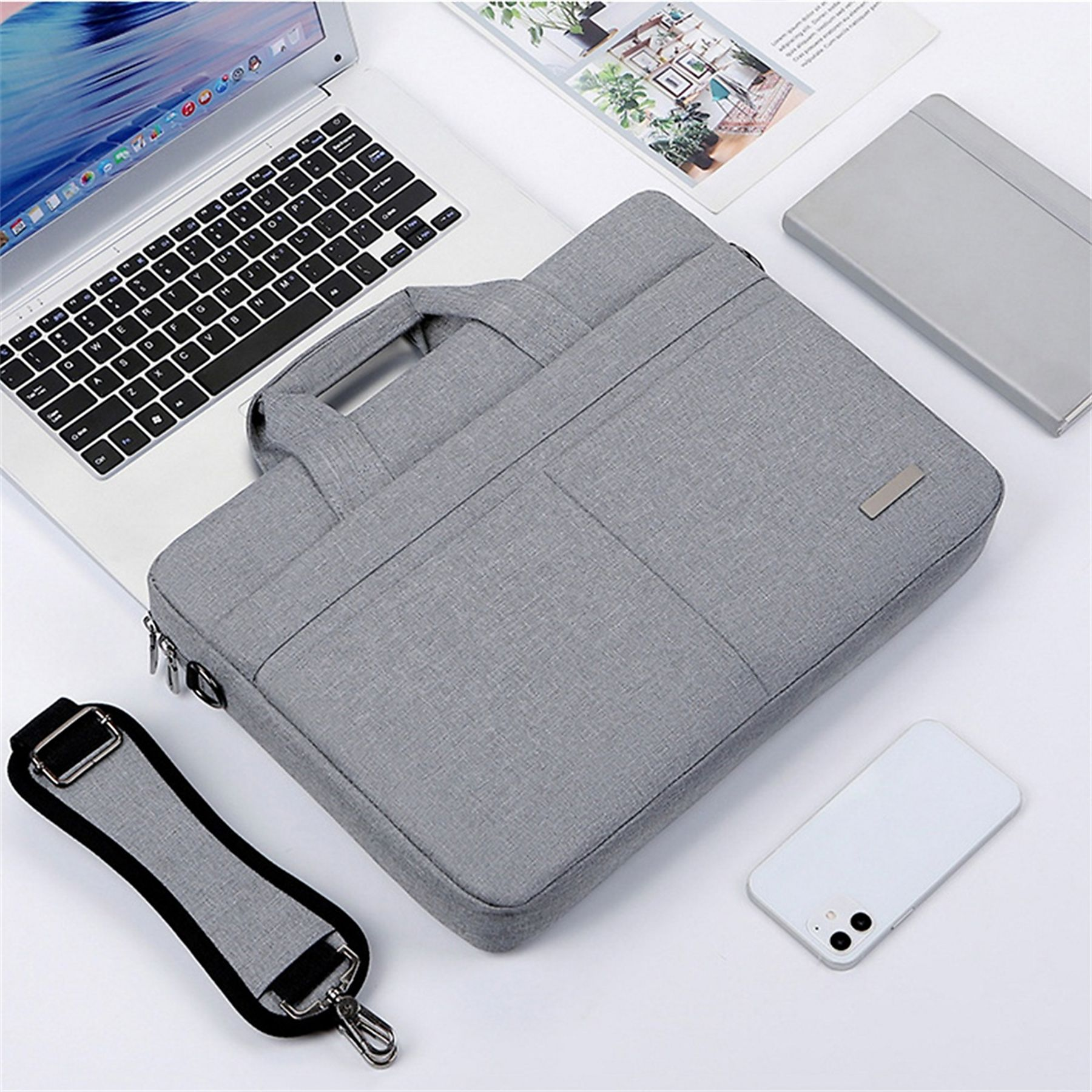 Graue Laptoptasche, Laptop, Telefon und Tablet liegen auf weißer Oberfläche. Die Tasche hat einen Griff und einen Gurt.