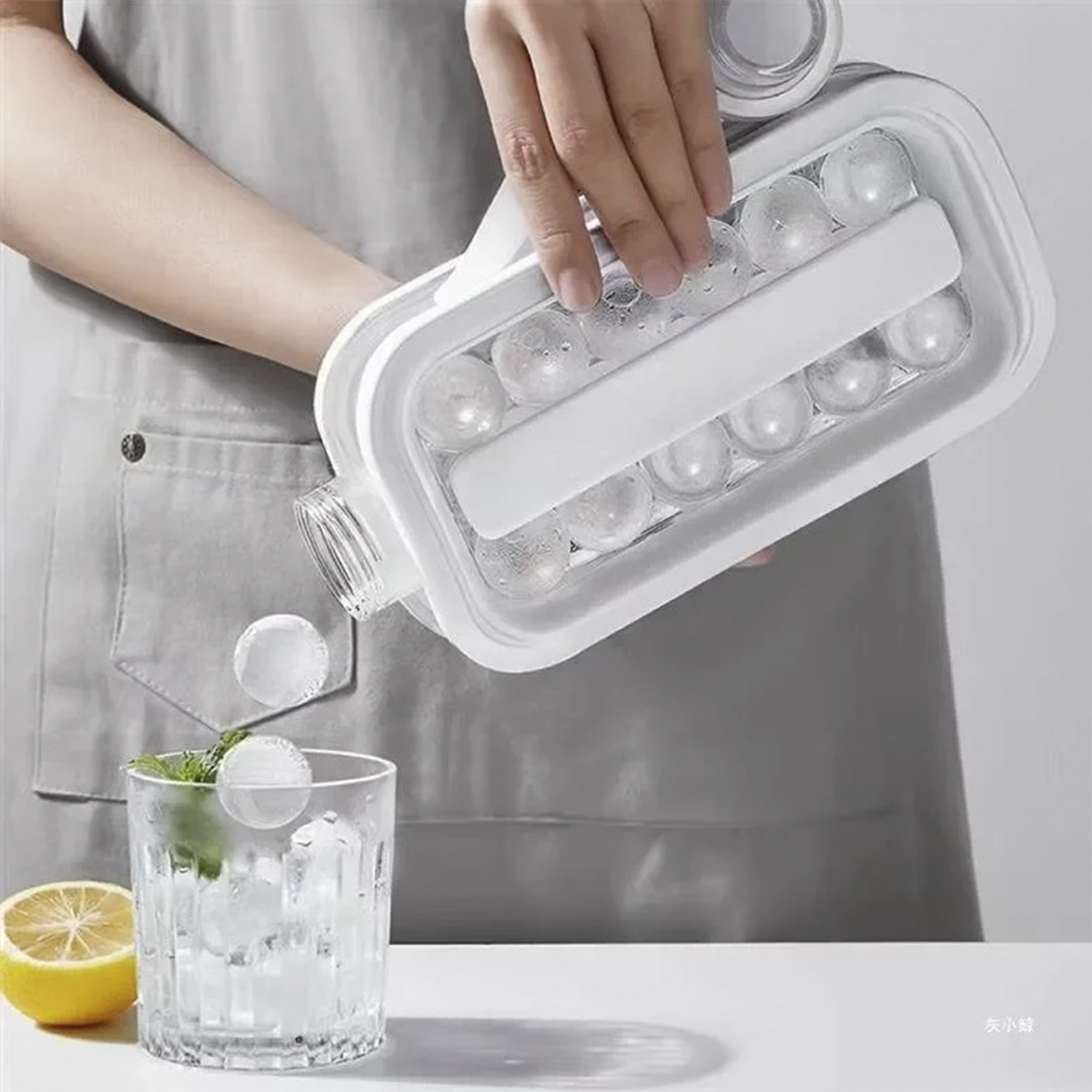 Una persona versa palline di ghiaccio da un contenitore bianco in un bicchiere con menta e limone. Lo sfondo è un grembiule grigio.