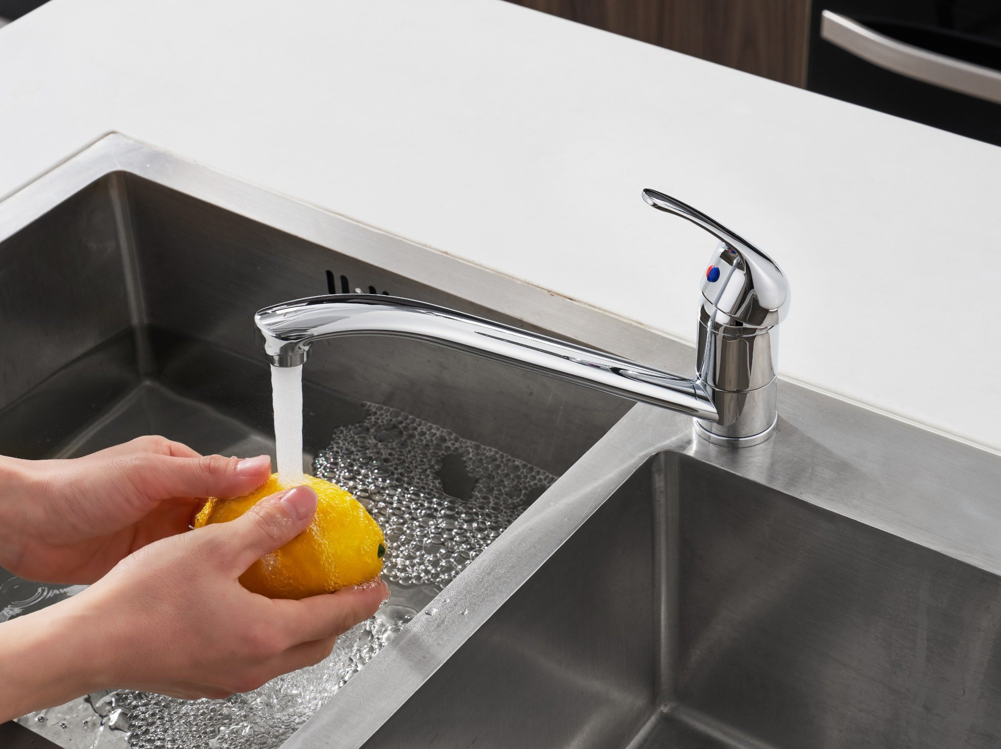 Hände waschen eine gelbe Zitrone unter fließendem Wasser aus einem verchromten Wasserhahn in einem Edelstahlbecken. Weiße Arbeitsplatte im Hintergrund.