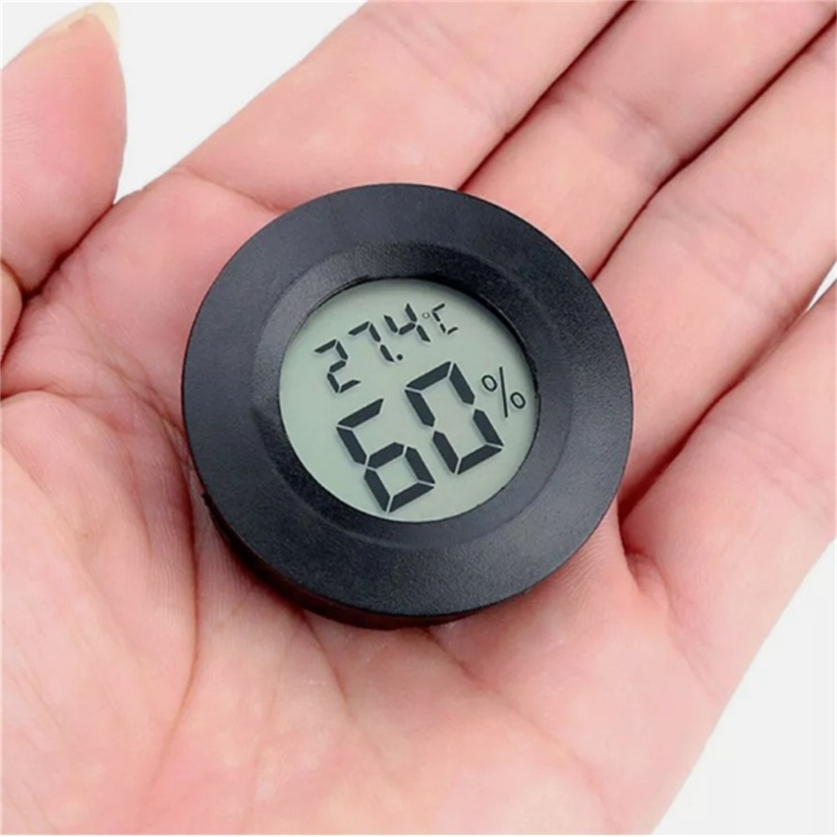 Ein schwarzes digitales Hygrometer, das 27,4 °C und 60 % anzeigt. Das Gerät wird in einer Handfläche gehalten.