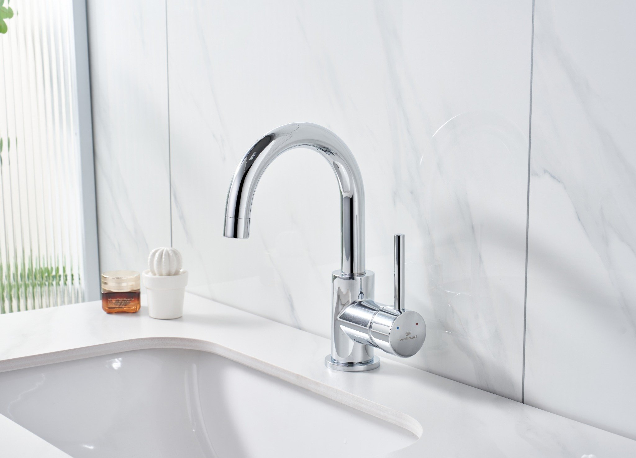 Glänzende Chromarmatur auf einer weißen Arbeitsplatte. Ein kleiner Kaktus und ein Glas stehen neben dem Waschbecken, mit einer Marmorwand im Hintergrund.