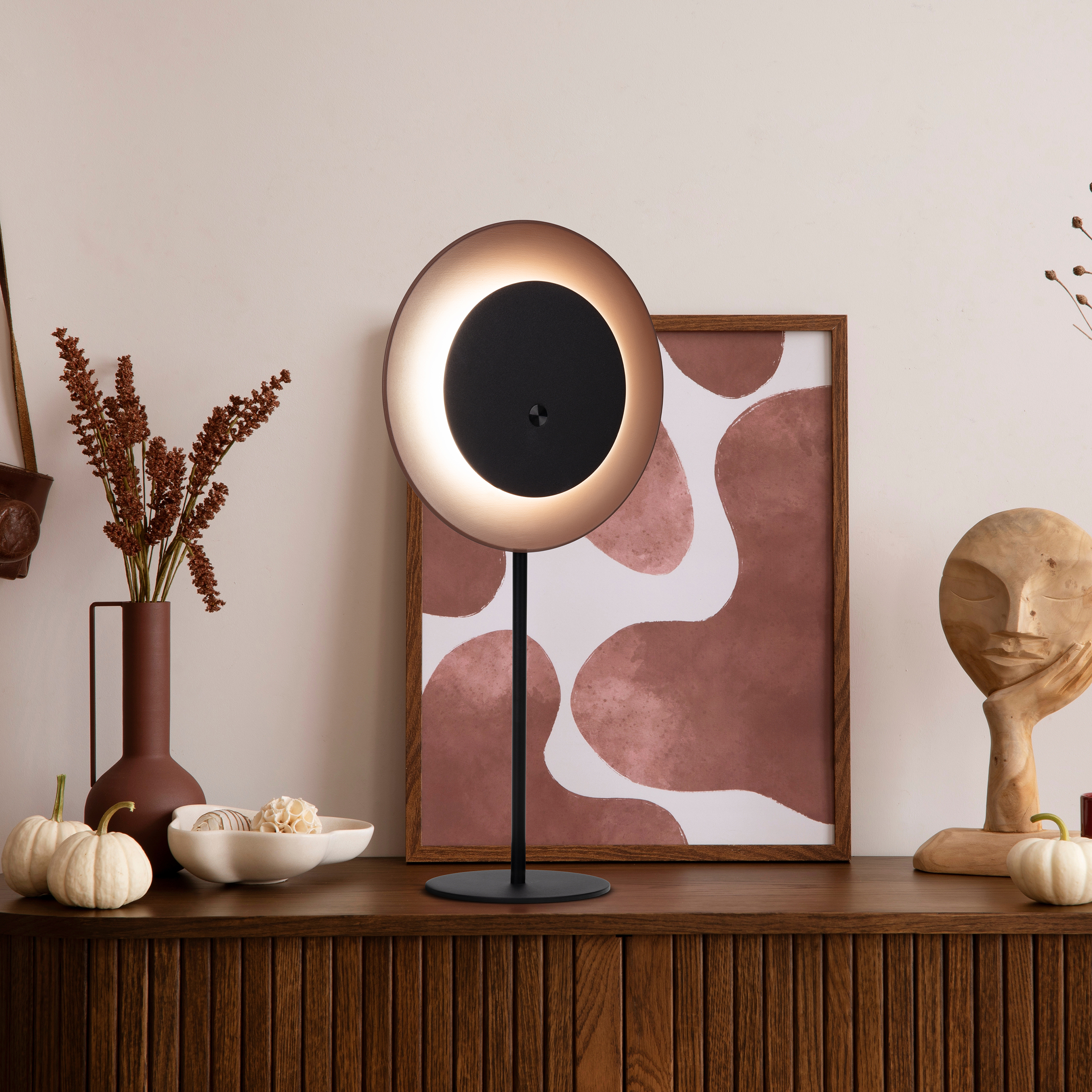 Ein Raum mit Lampe, Bilderrahmen, Vase, Kürbissen und einer Holzskulptur auf einem braunen Schrank.