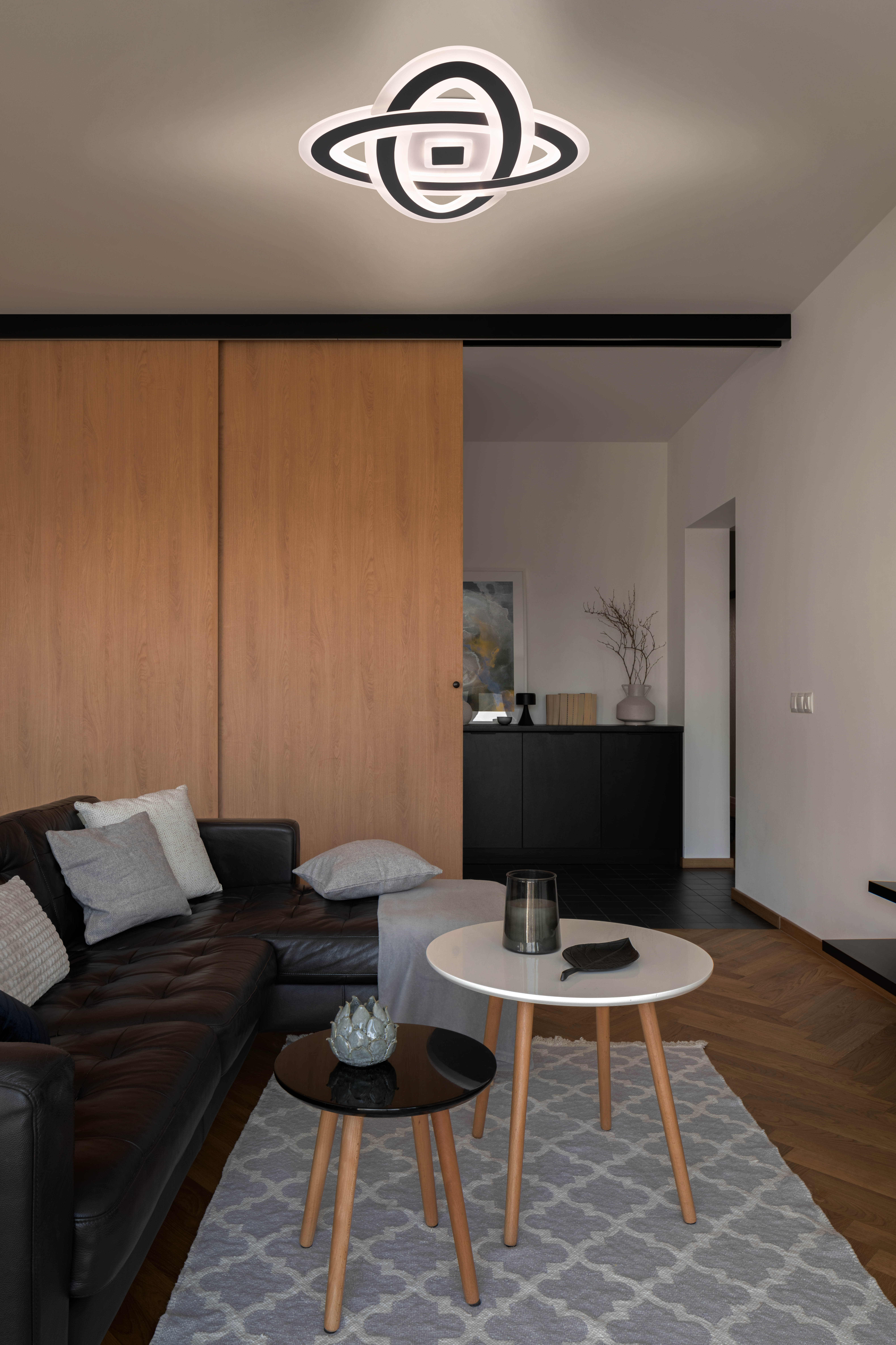 Wohnzimmer mit schwarzem Ledersofa, Holztüren und weiß-schwarzer Deckenleuchte. Zwei kleine Tische mit grauem Teppich.