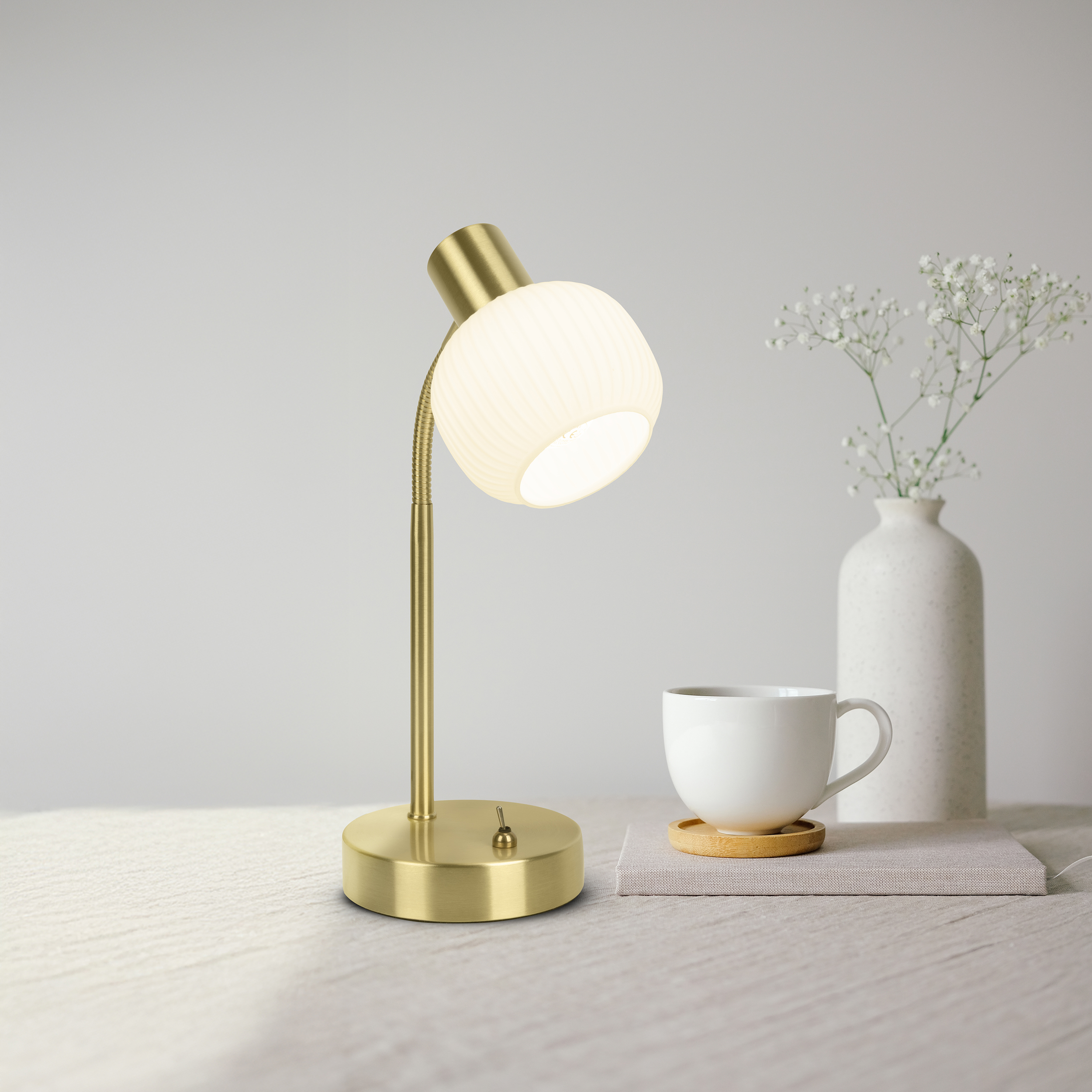 Eine Messing-Schreibtischlampe mit beleuchtetem Glasschirm. Eine weiße Kaffeetasse und eine Vase mit Blumen sind ebenfalls vorhanden.