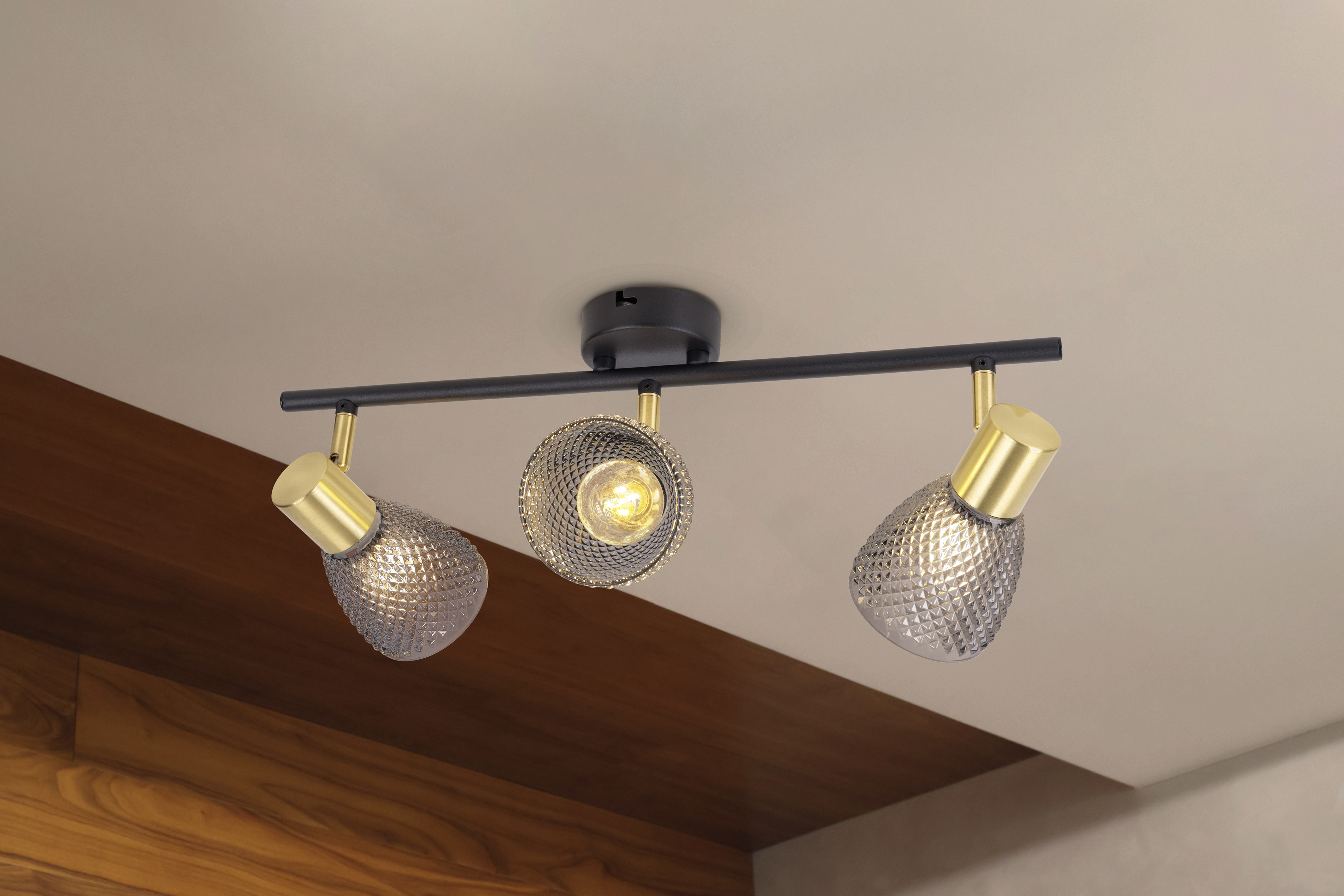 Deckenleuchte mit drei verstellbaren Köpfen. Jeder Kopf hat einen goldfarbenen Sockel und einen strukturierten Glasschirm. Hintergrund ist beige und Holz.