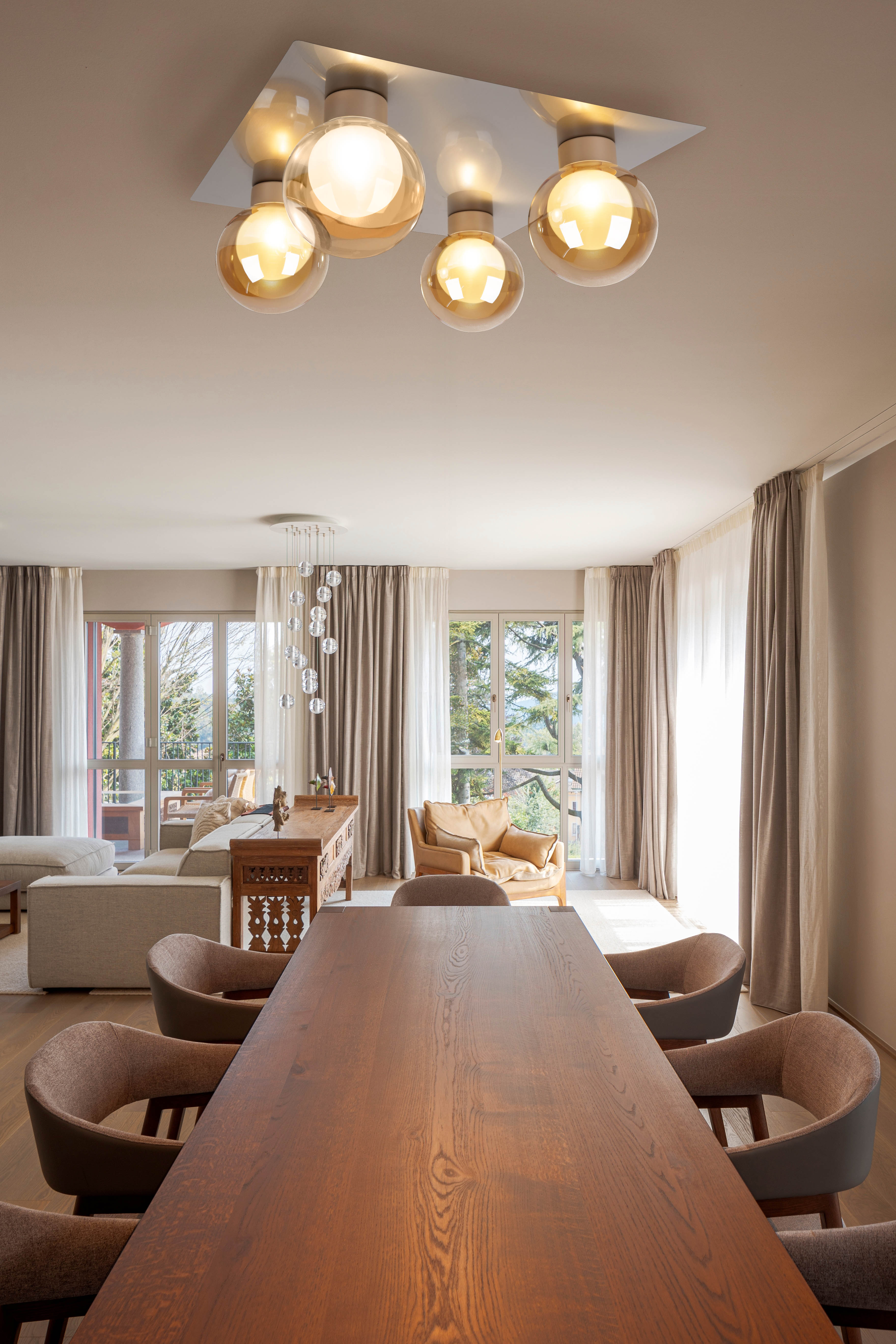 Esszimmer mit großem Holztisch, Stühlen und Leuchten. Fenster mit Vorhängen. Sofa und Sessel sind sichtbar.
